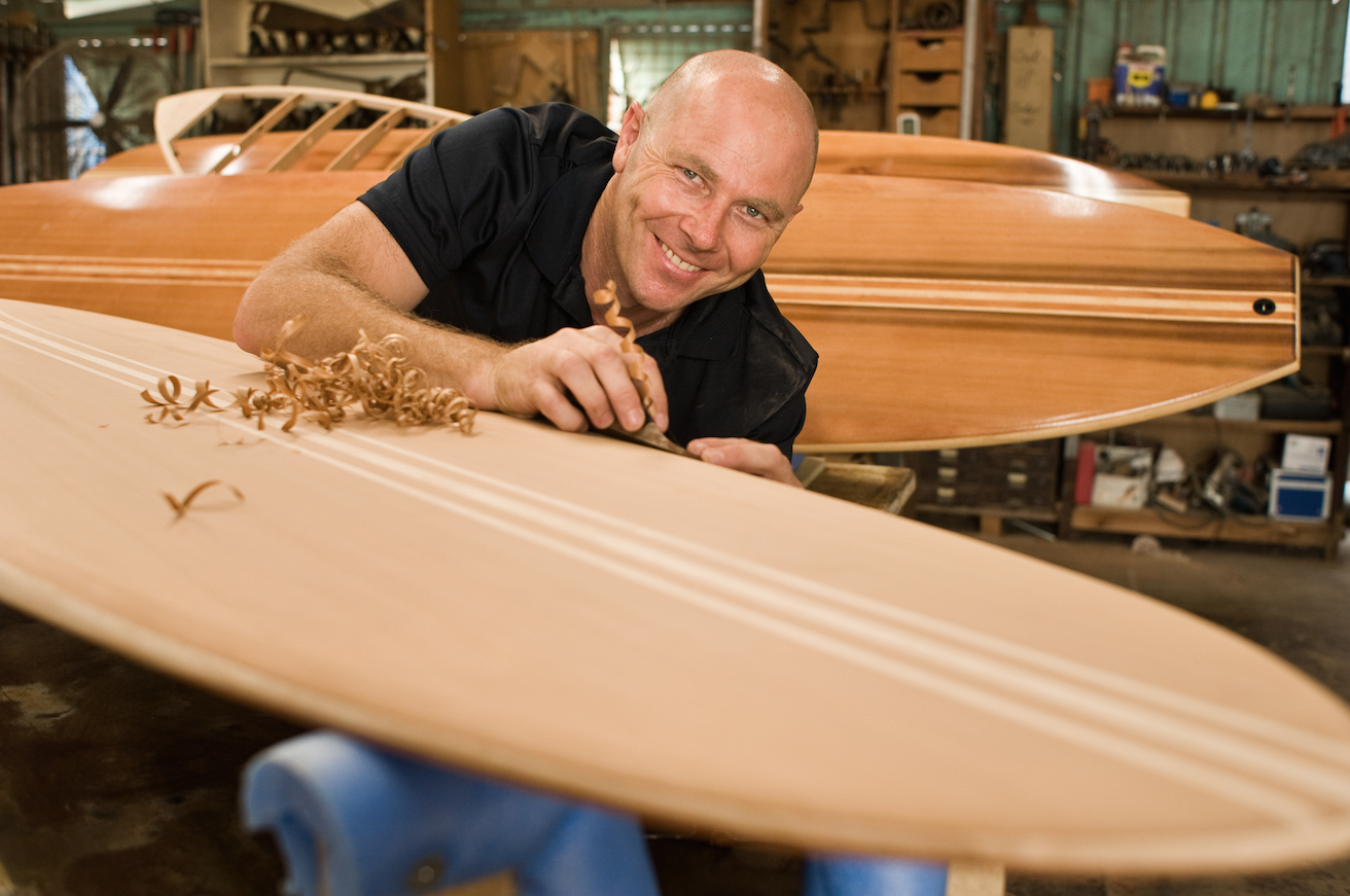 A man smiling while working on a wooden surfboard in a woodworking shop.