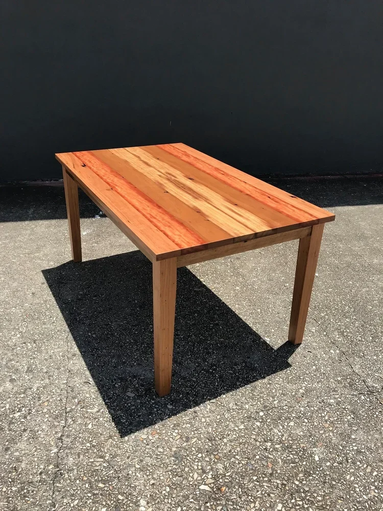 Wooden table with a colorful wood top on an asphalt surface outside, casting a shadow.