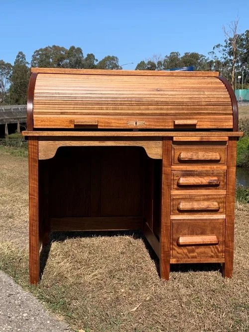 Wooden roll-top desk with four drawers on the right side, placed outdoors on grass with trees and blue sky in background.