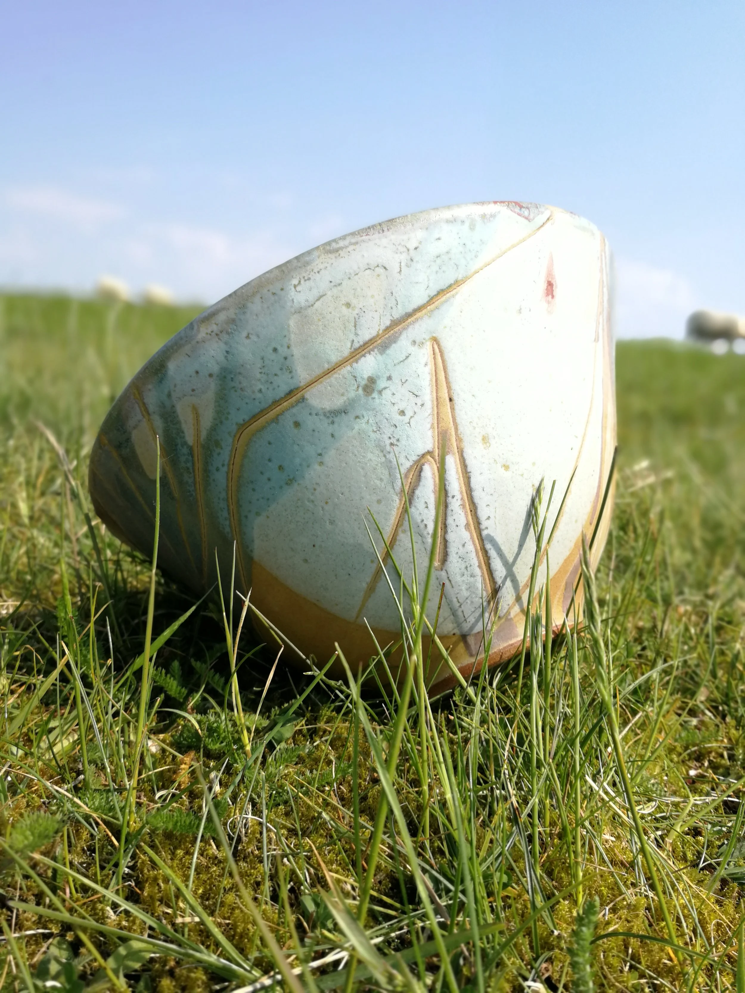 A large decorative stone with a smooth surface and painted abstract patterns resting on green grass in a sunny outdoor field with a clear blue sky in the background.