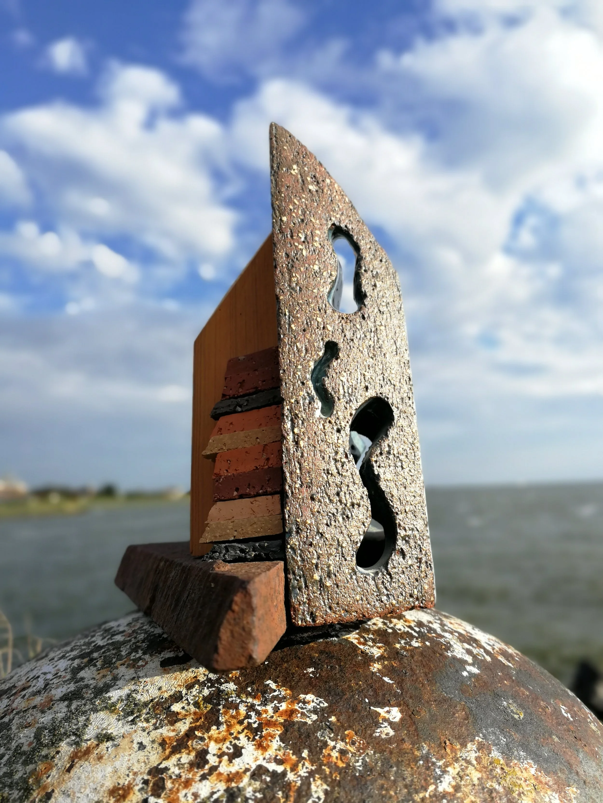 A close-up photo of a metal and wooden sculpture resembling a house, with a background of cloudy sky and water.