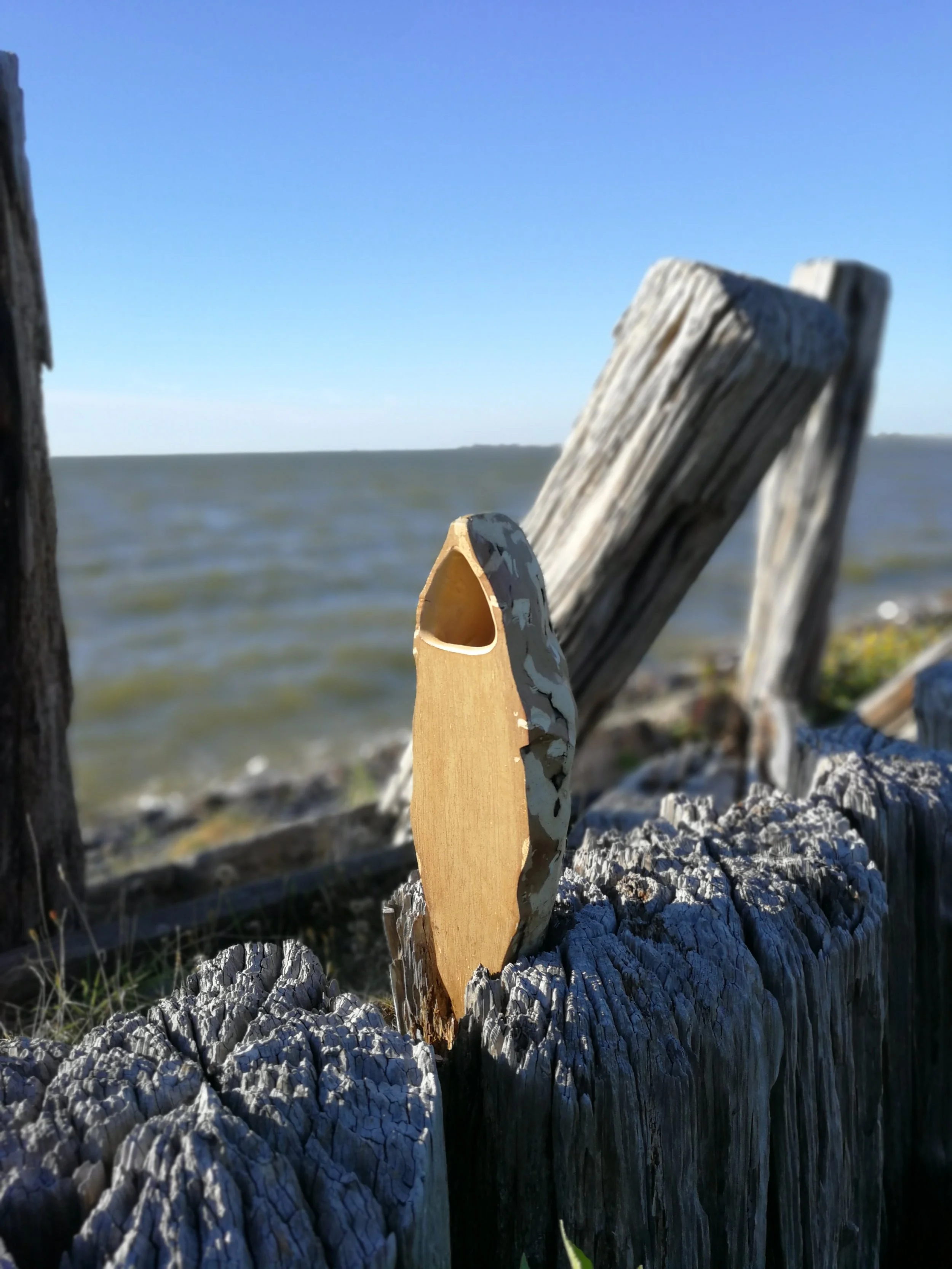 Close-up photo of a weathered wooden post near water with a small wooden bird-shaped carving attached, against a background of a calm body of water and a clear blue sky.