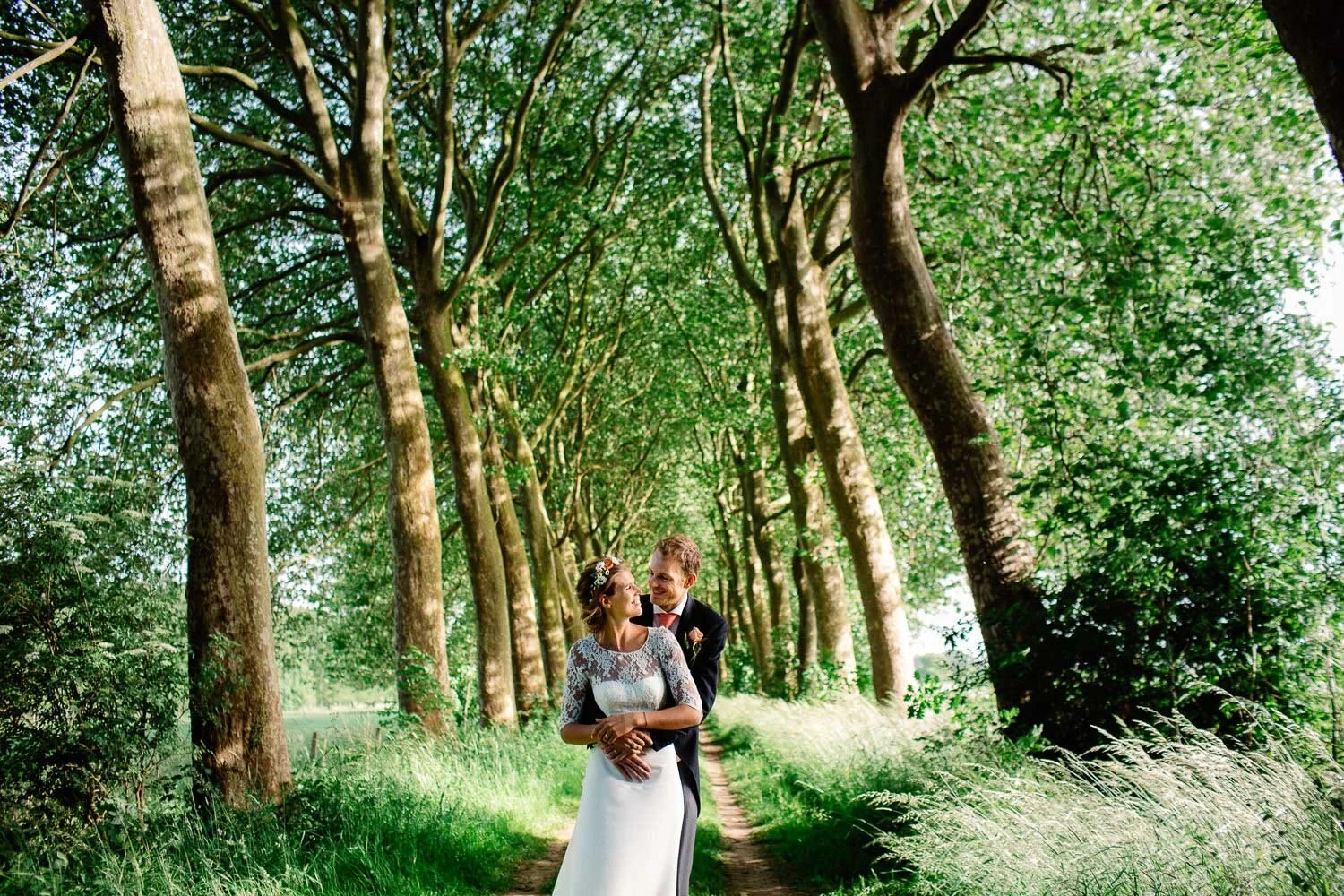 A bride and groom standing closely together on a tree-lined path, looking at each other affectionately in a lush green park.