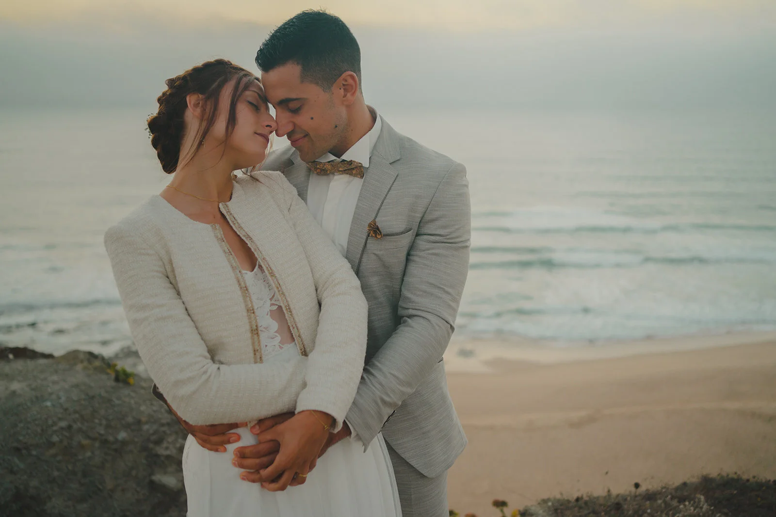 A couple dressed in wedding attire embracing on a beach at sunset, with the ocean in the background.