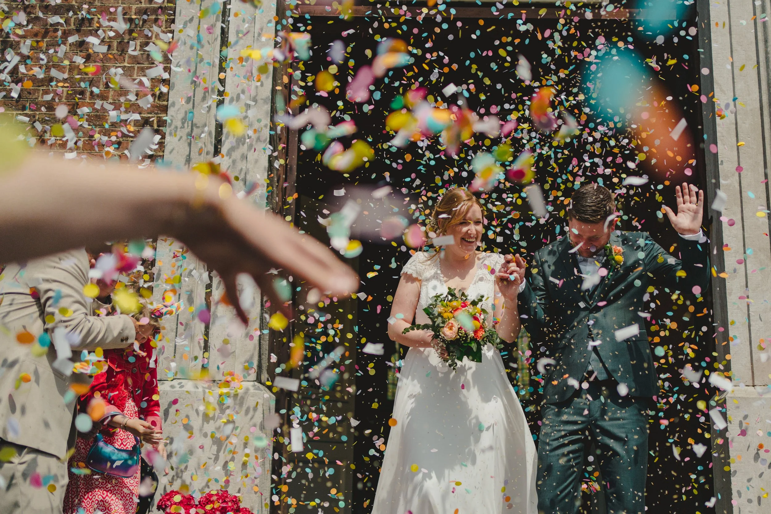 A newlywed couple exiting a building under a shower of colorful confetti, smiling and holding hands, with wedding attire and a bouquet of flowers.