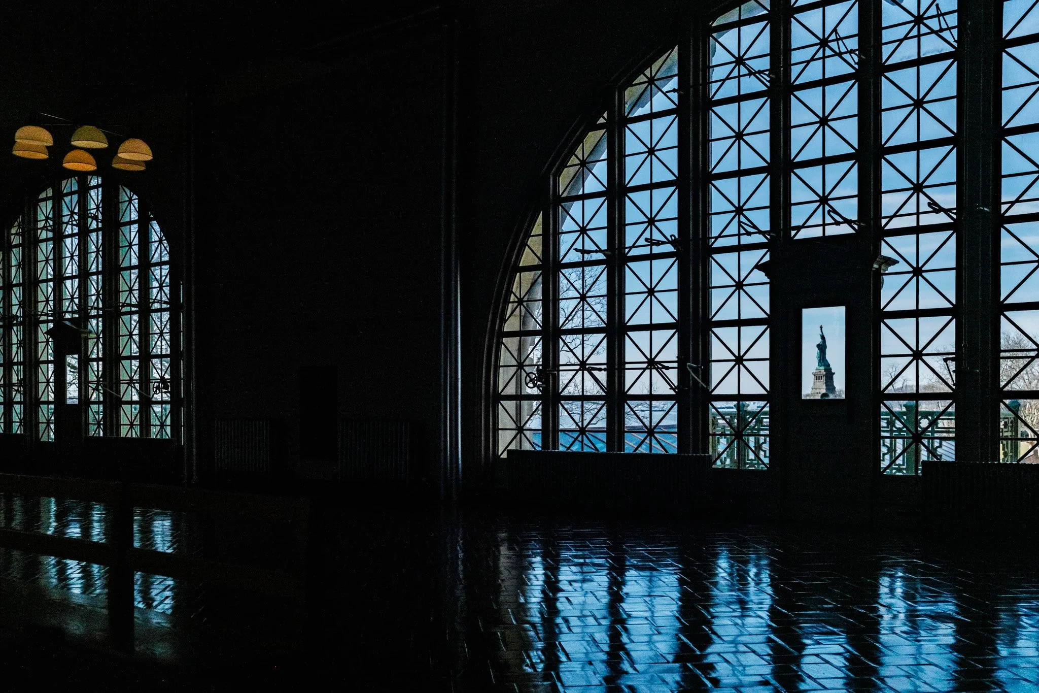 Interior view of a dark building with large arched windows showing the Statue of Liberty in the distance against a blue sky.