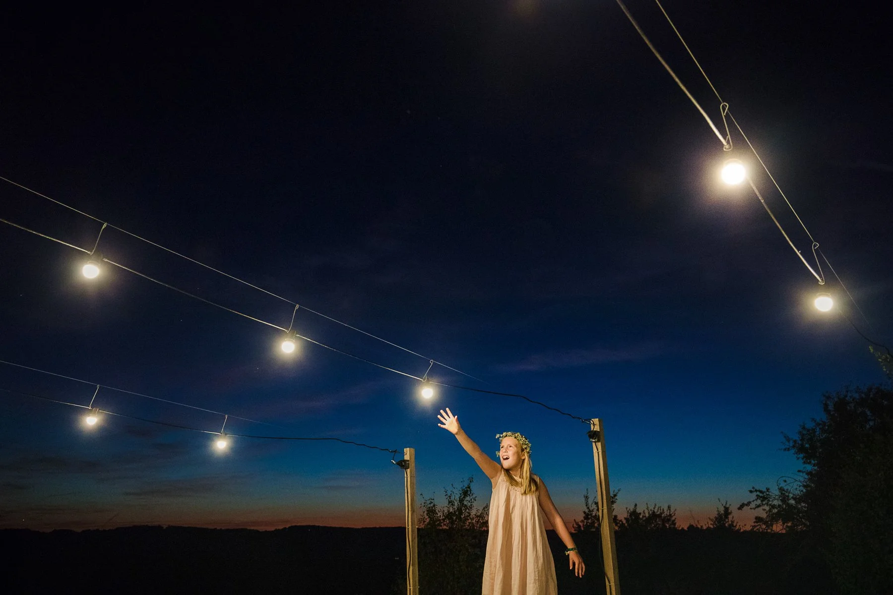 A young woman in a beige dress and floral crown standing outdoors at night, reaching up towards string lights hung above her against a darkening sky.