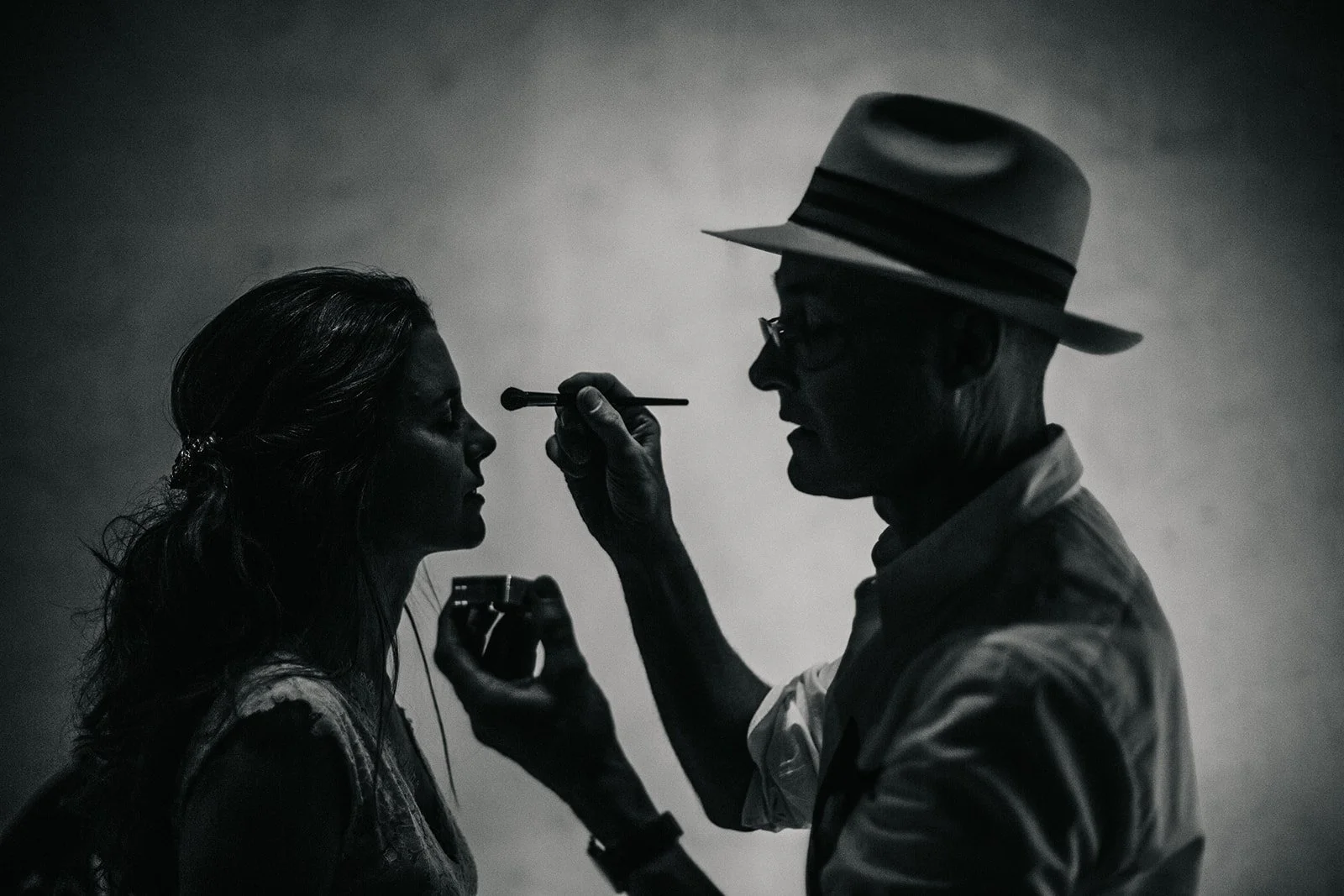 A man applying makeup to a woman, shown in silhouette, with a plain background.