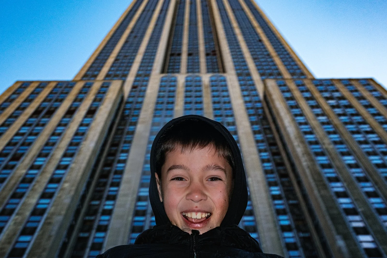 A young boy with short brown hair smiling in front of the towering Empire State Building during daytime.
