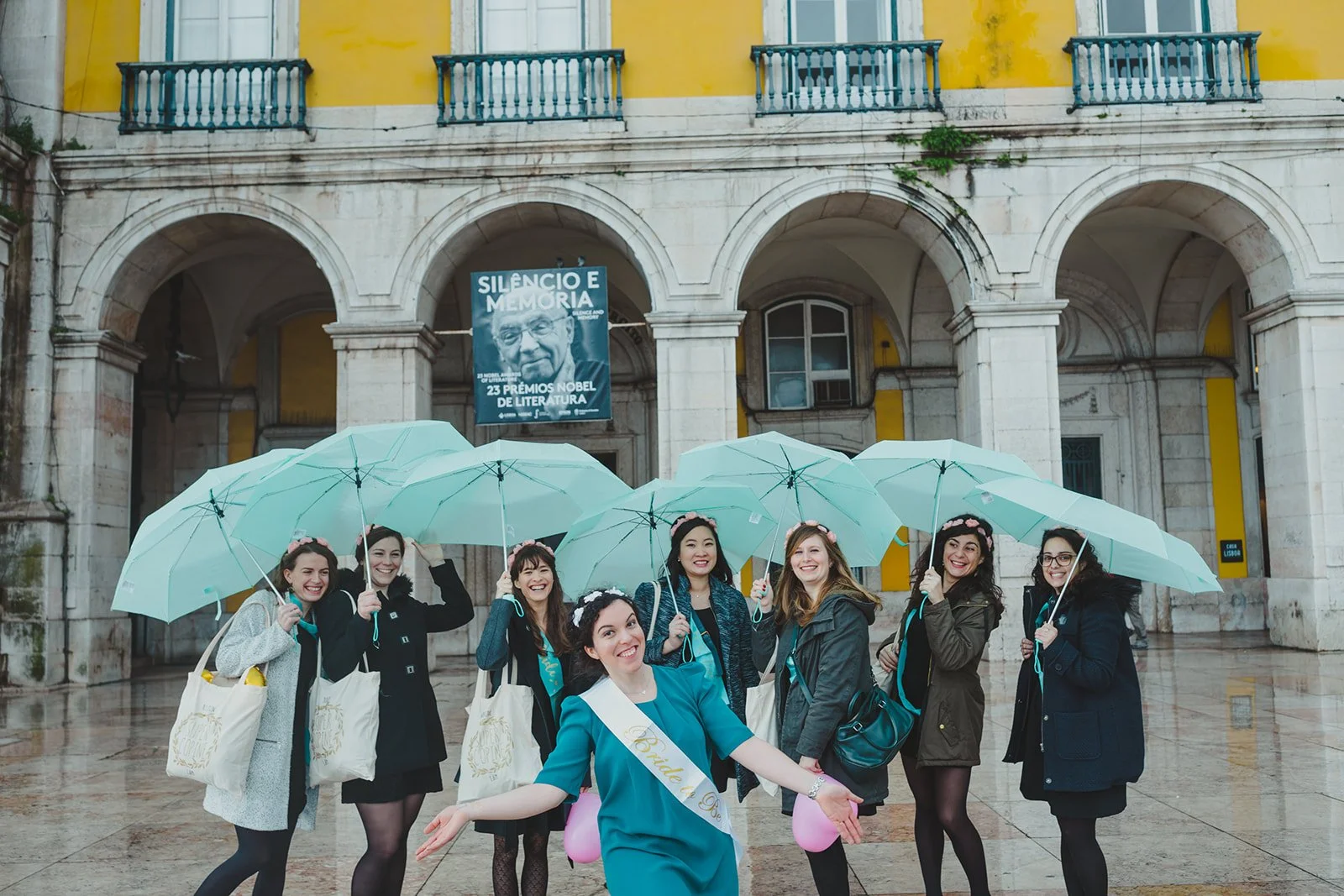 Group of women celebrating a wedding outdoors in the rain, holding turquoise umbrellas, with a woman wearing a sash and tiara, smiling.