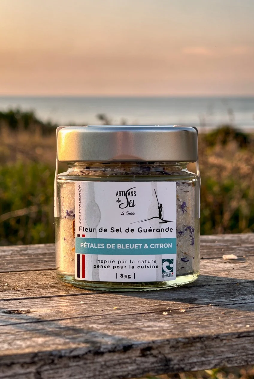 Pot de fleur de sel de Guérande aromatisée aux pétales de bleuet et citron, posé sur une table en bois devant une mer au coucher du soleil.