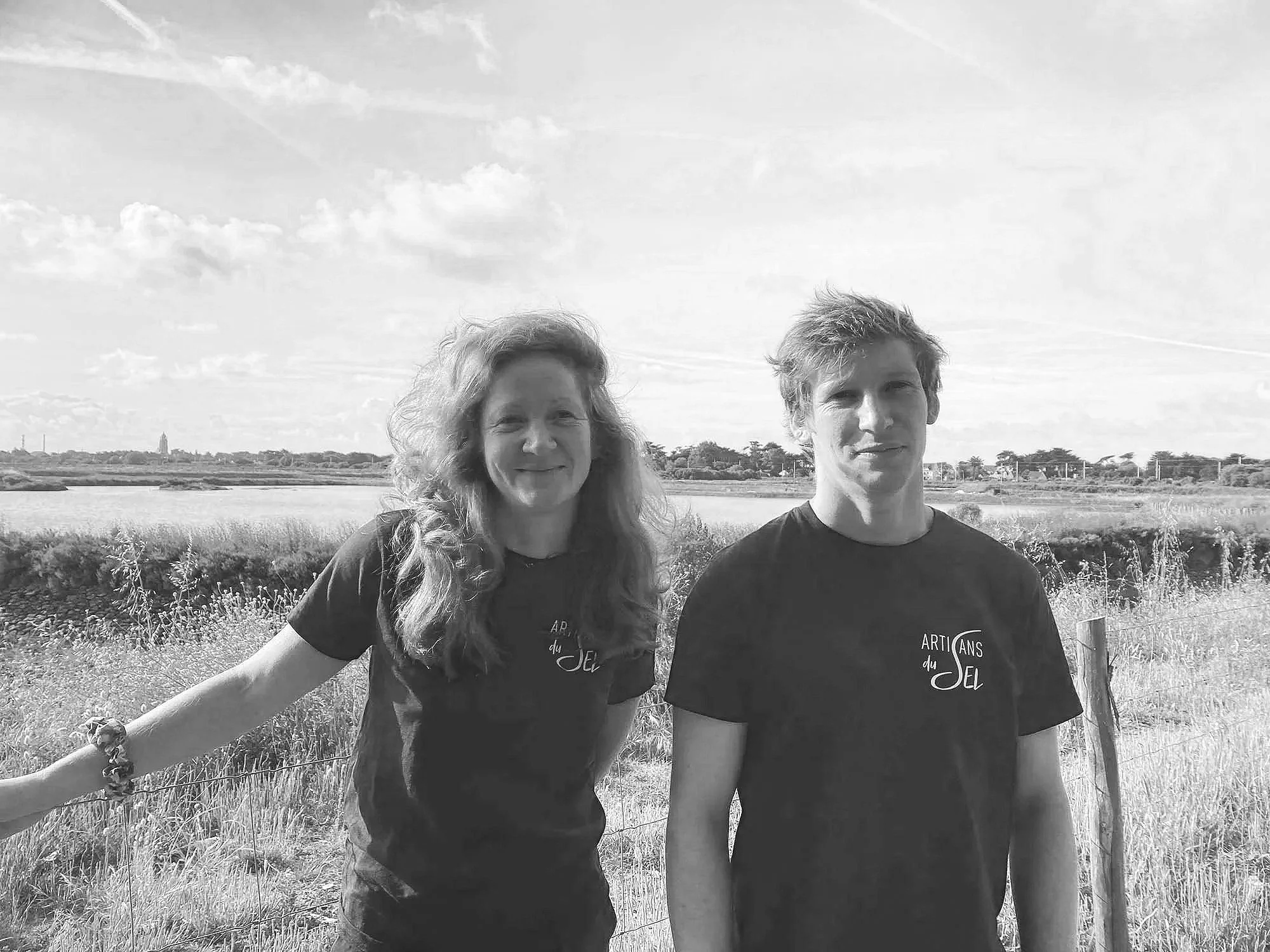 Deux personnes, un homme et une femme, posent ensemble devant un paysage rural avec étangs et arbres sous un ciel partiellement nuageux.