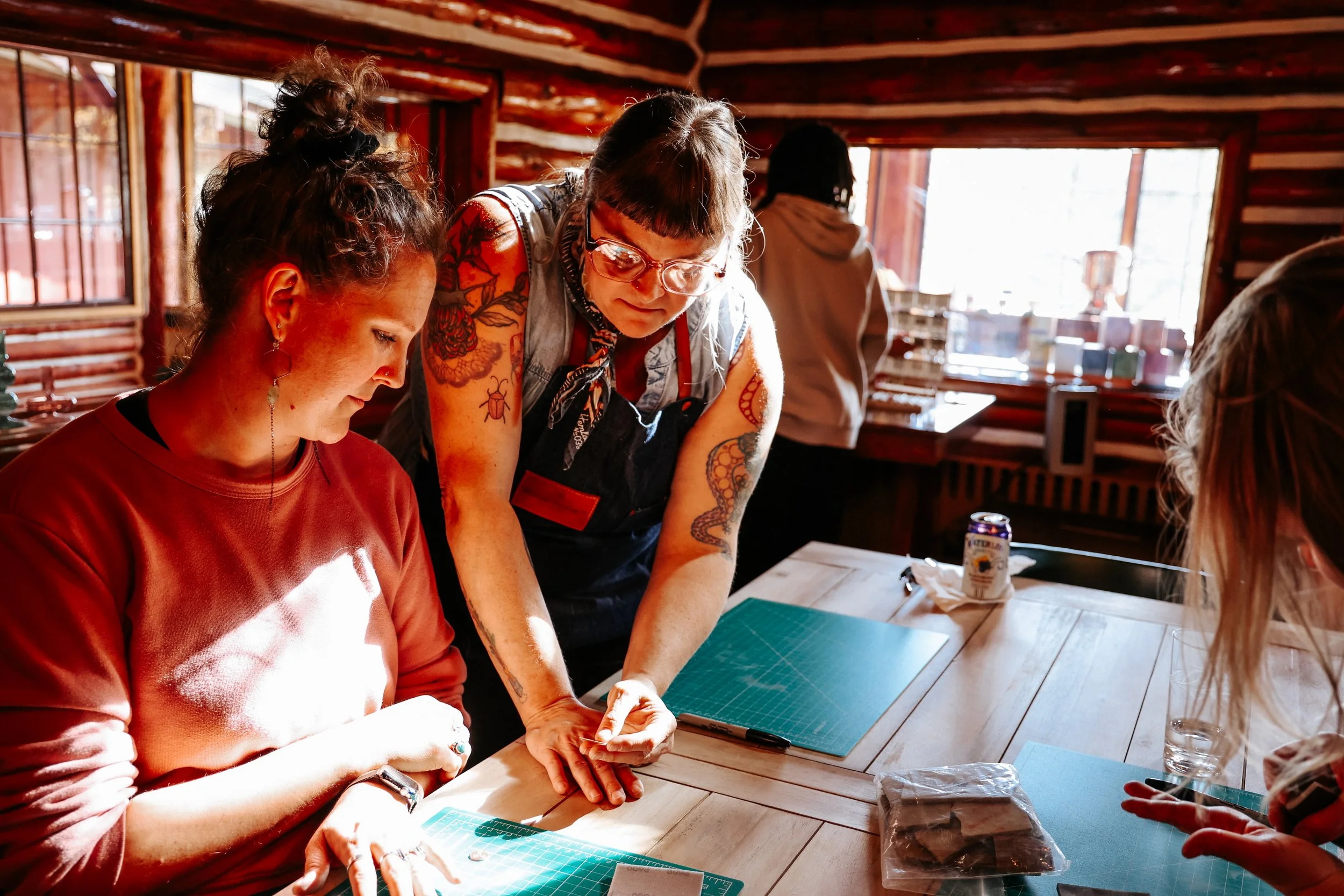 Two women sit at a table; one is helping the other create beautiful metal jewelry.
