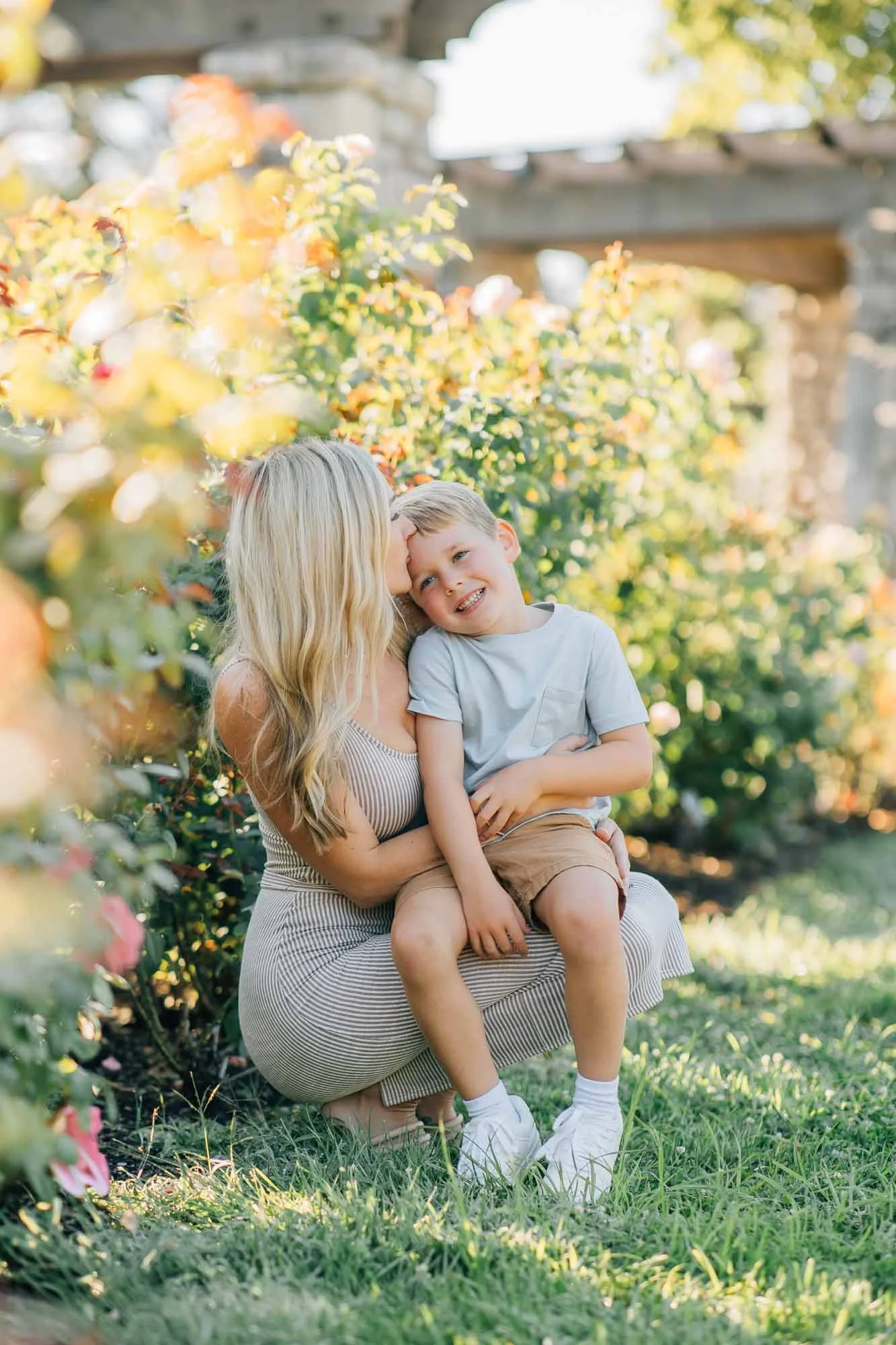 Mother kissing sons forehead while kneeling in rose garden, Kansas City family photographer