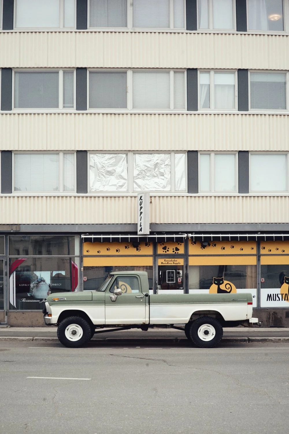 Retro pickup-auto pysäköitynä kaupunkikadulle kerrostalon ja kuppilan eteen. Rakennuksen ikkunat ja kuppilan keltainen mainoskyltti näkyvät taustalla.