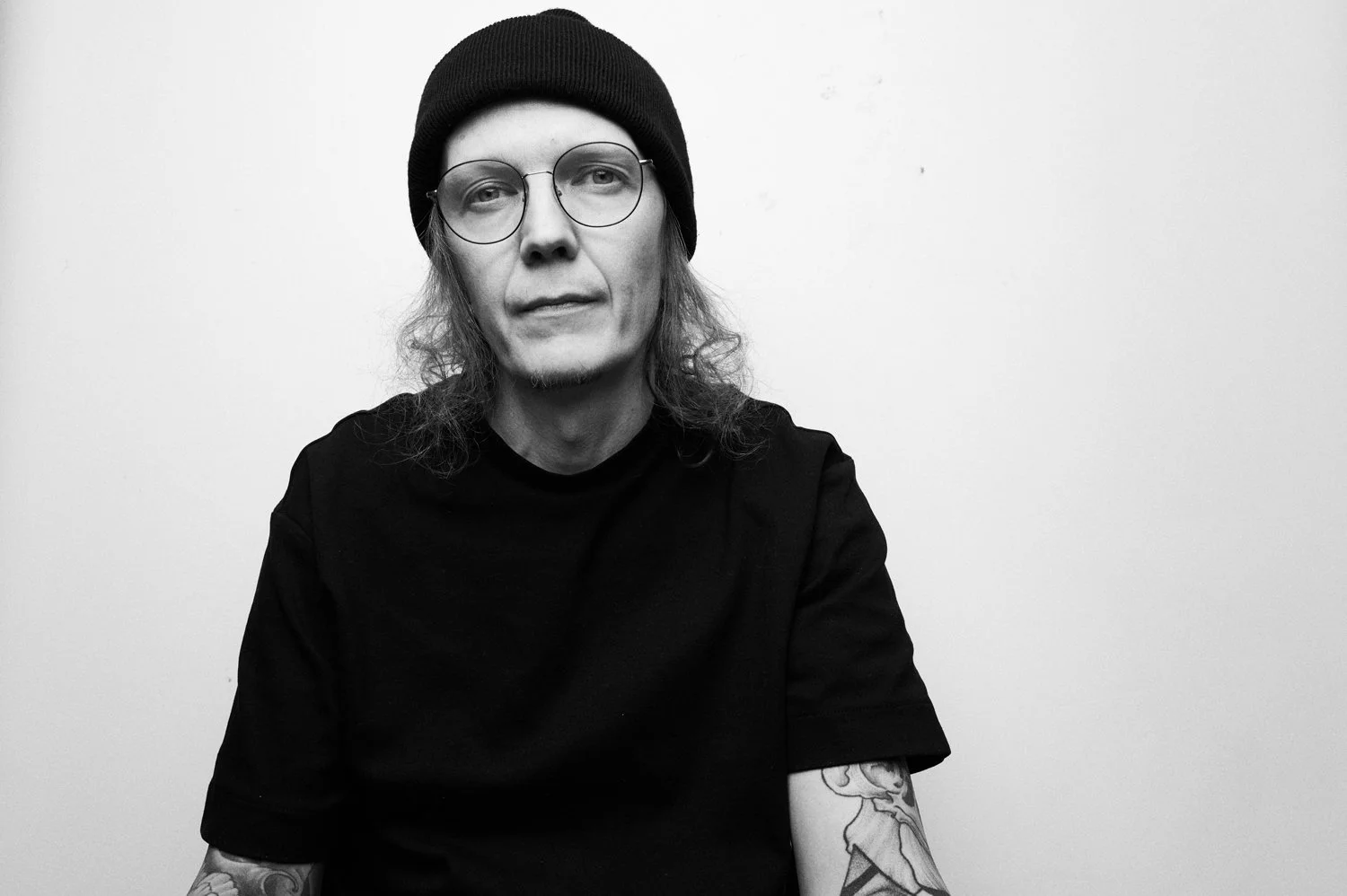 Mustavalkovalokuva miespuolisesta henkilöstä, jolla on pitkät hiukset, lasit ja tatuointeja, istuu tai seisoo vaaleanharmaalla taustalla, pukeutuneena mustaan t-paitaan ja pipo.