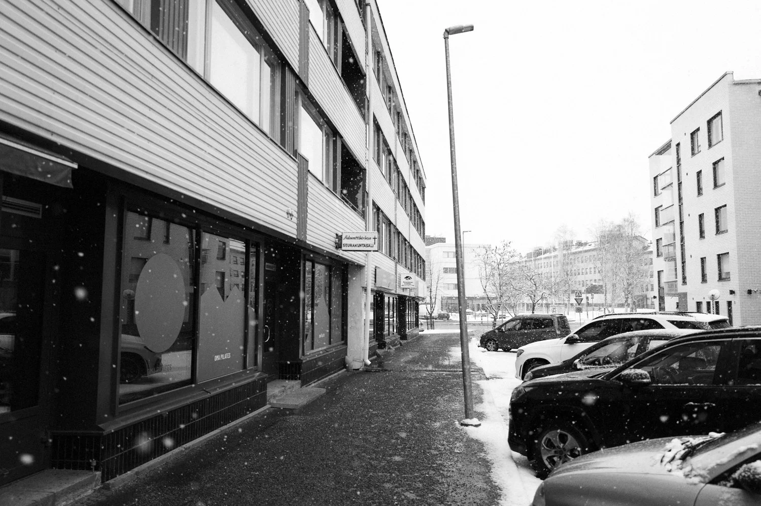 Mustavalkoinen valokuva kadusta talvella, jossa on lunta satamassa. Kadun varrella on rakennuksia ja pysäköityjä autoja. Rakennuksissa liiketiloja ja kadulla katulamppu.