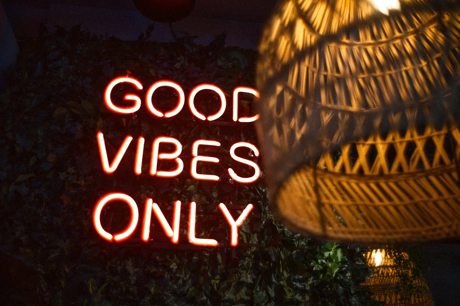Neon-kyltti "Good Vibes Only" tummalla taustalla ja rottinkilamppu etualalla.