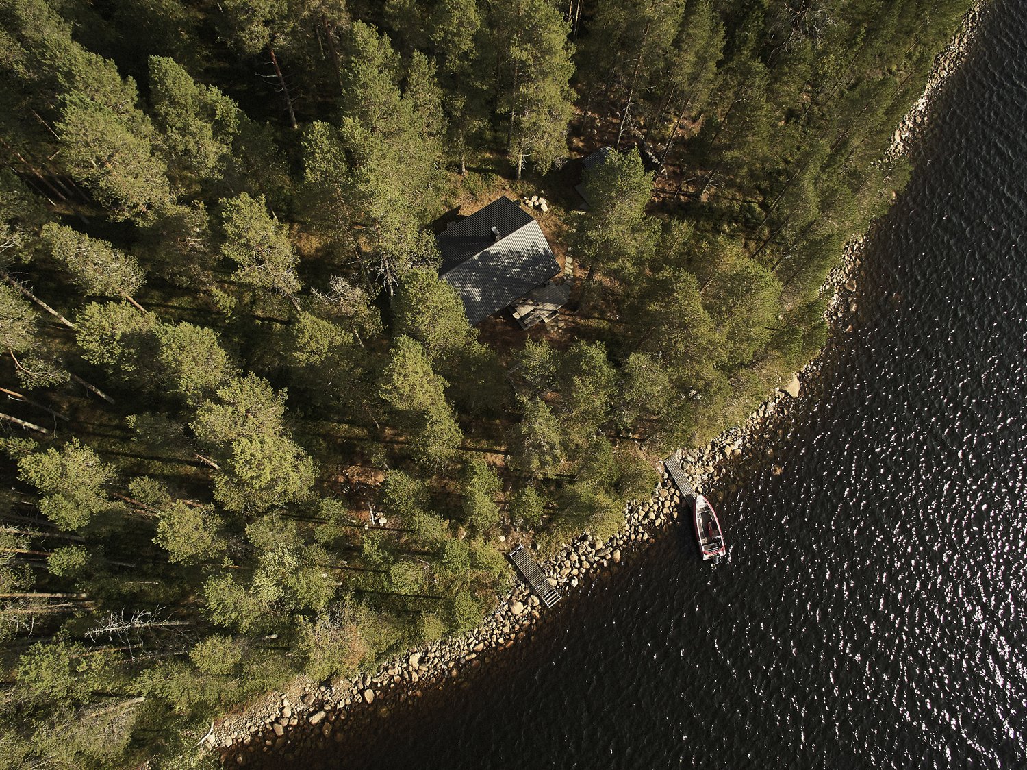 Ilmakuva metsäisestä alueesta, jossa on mökki ja laituri. Laiturissa on vene, ja vieressä on järvi. Puita näkyy ympäriinsä.