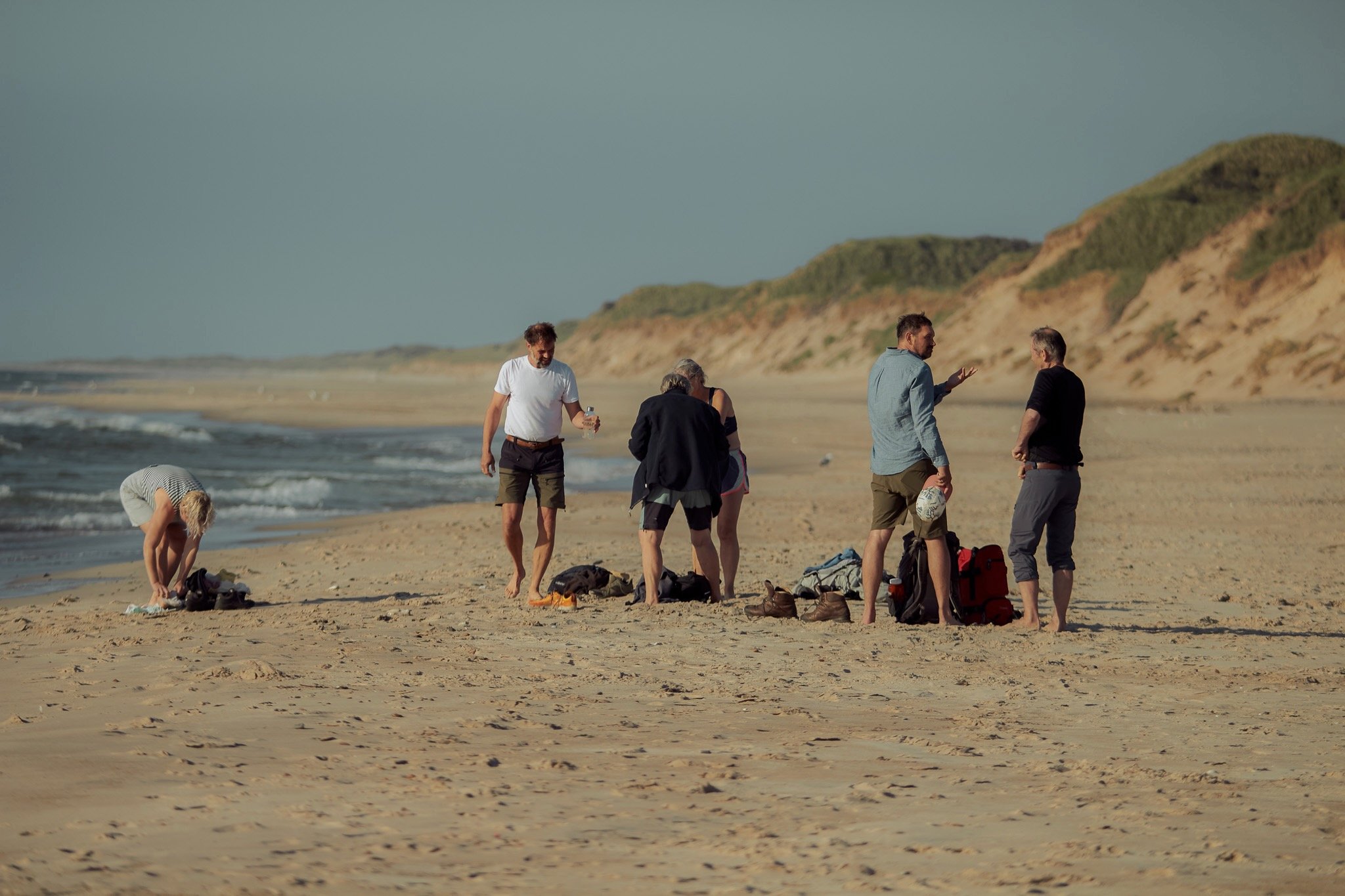 En gruppe mennesker på en strand, nogle står og snakker, en person bøjer sig ned for at pakke udstyr, alle omgivet af sand og klitter i baggrunden.