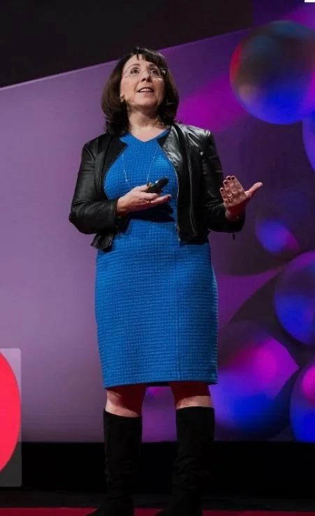 Una donna che tiene un microfono e parla durante una presentazione o conferenza in un palco, con uno sfondo colorato e decorato con cerchi colorati