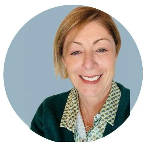 Foto di una donna sorridente con capelli corti, abbigliata con una camicia verde e un maglione scuro, su sfondo blu chiaro.