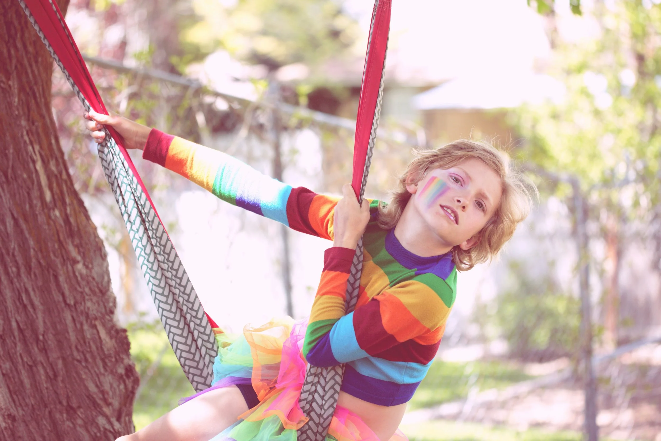 Una bambina con trucco arcobaleno sul volto, indossa abiti colorati e un tutù a strisce arcobaleno, si arrampica su un albero in un parco soleggiato.