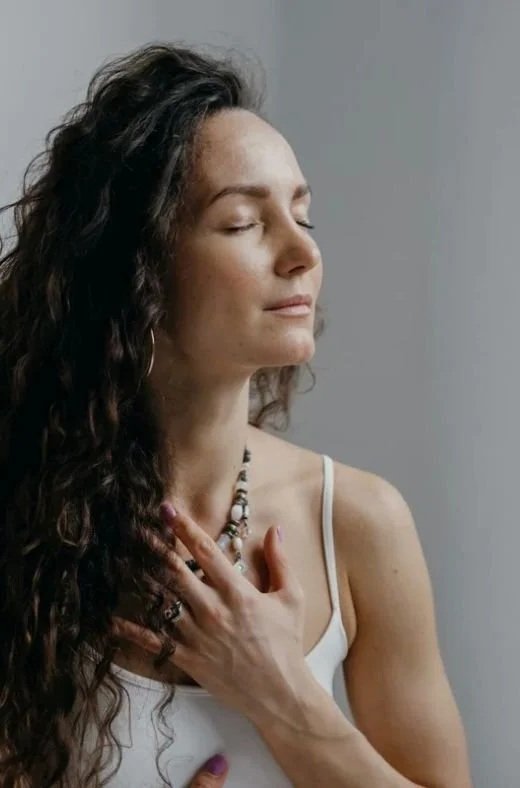 Donna con occhi chiusi, capelli ricci, indossa una canotta bianca e accessori, con la mano sul petto, davanti a uno sfondo grigio.