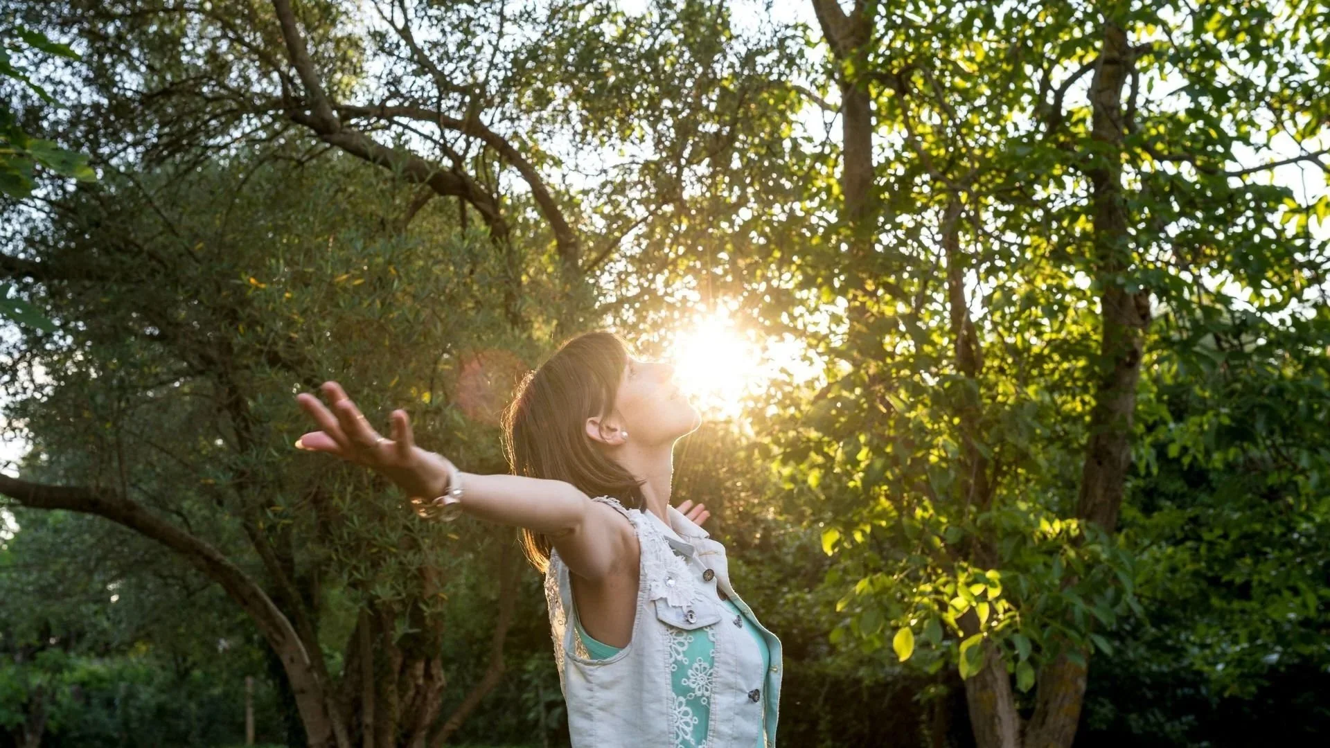 Una donna con braccia aperte e volto rivolto verso il sole tra gli alberi di un bosco verde in una giornata soleggiata.