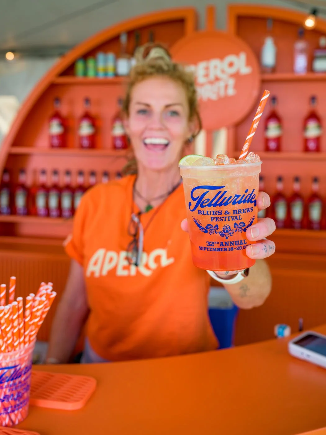 A smiling representative from Aperol Spritz holds up a festival glass of the classic cocktail, Aperol Spritz, at the festival in a cup with the logo on it.