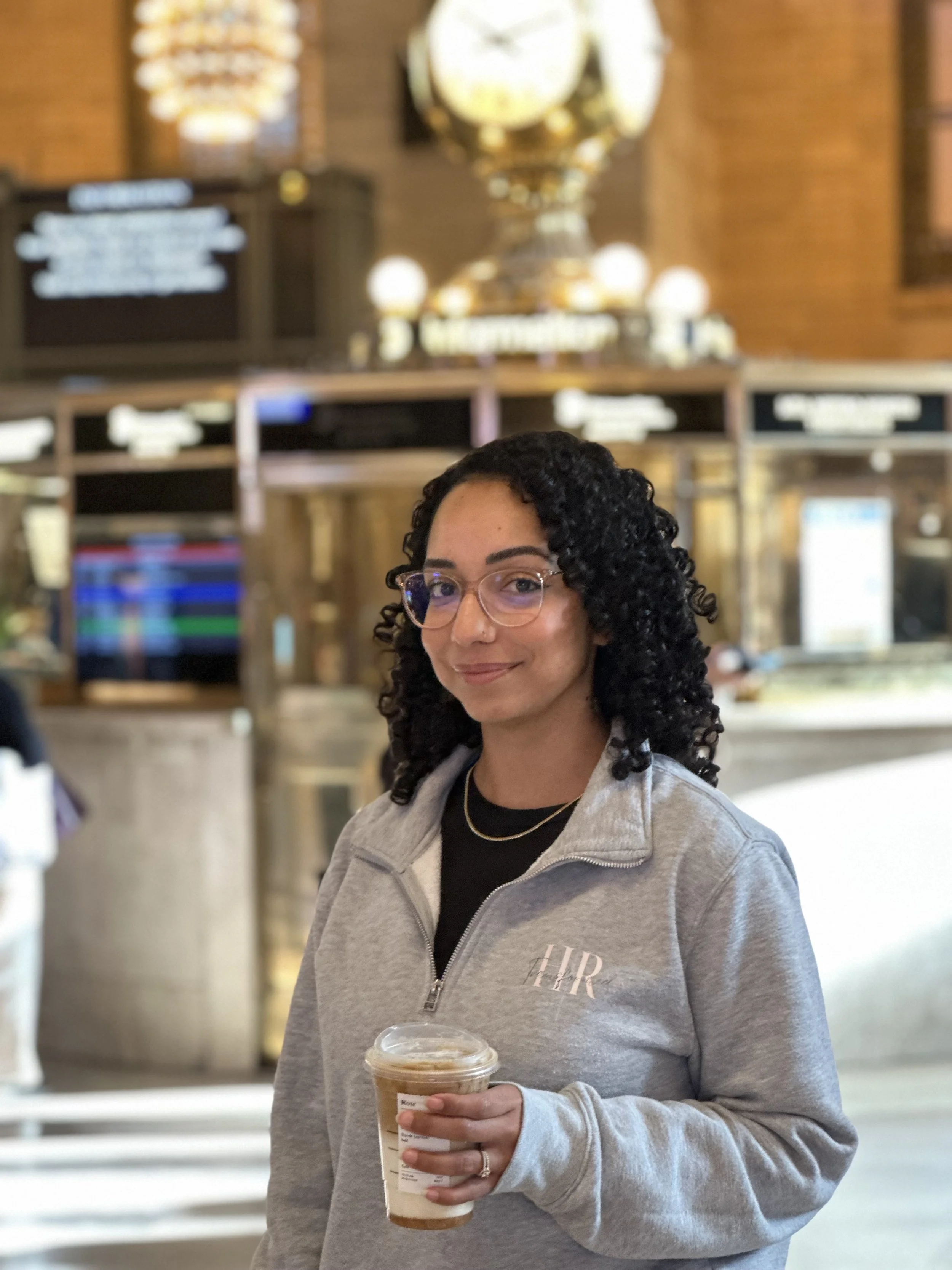 A woman with curly dark hair wearing glasses and a gray zip-up jacket holding iced coffee inside a coffee shop or cafe.