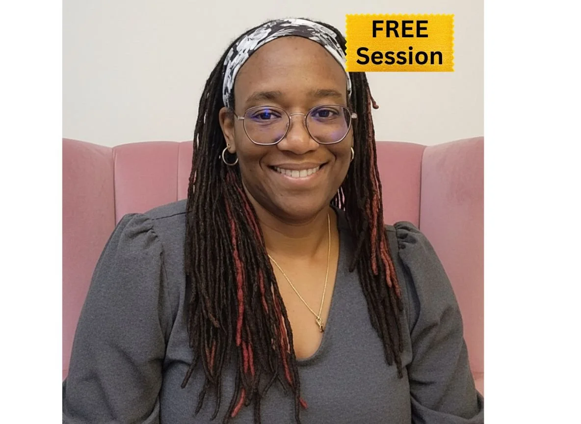 A woman with glasses and braids smiling, sitting on a pink couch, wearing a gray top and gold jewelry.