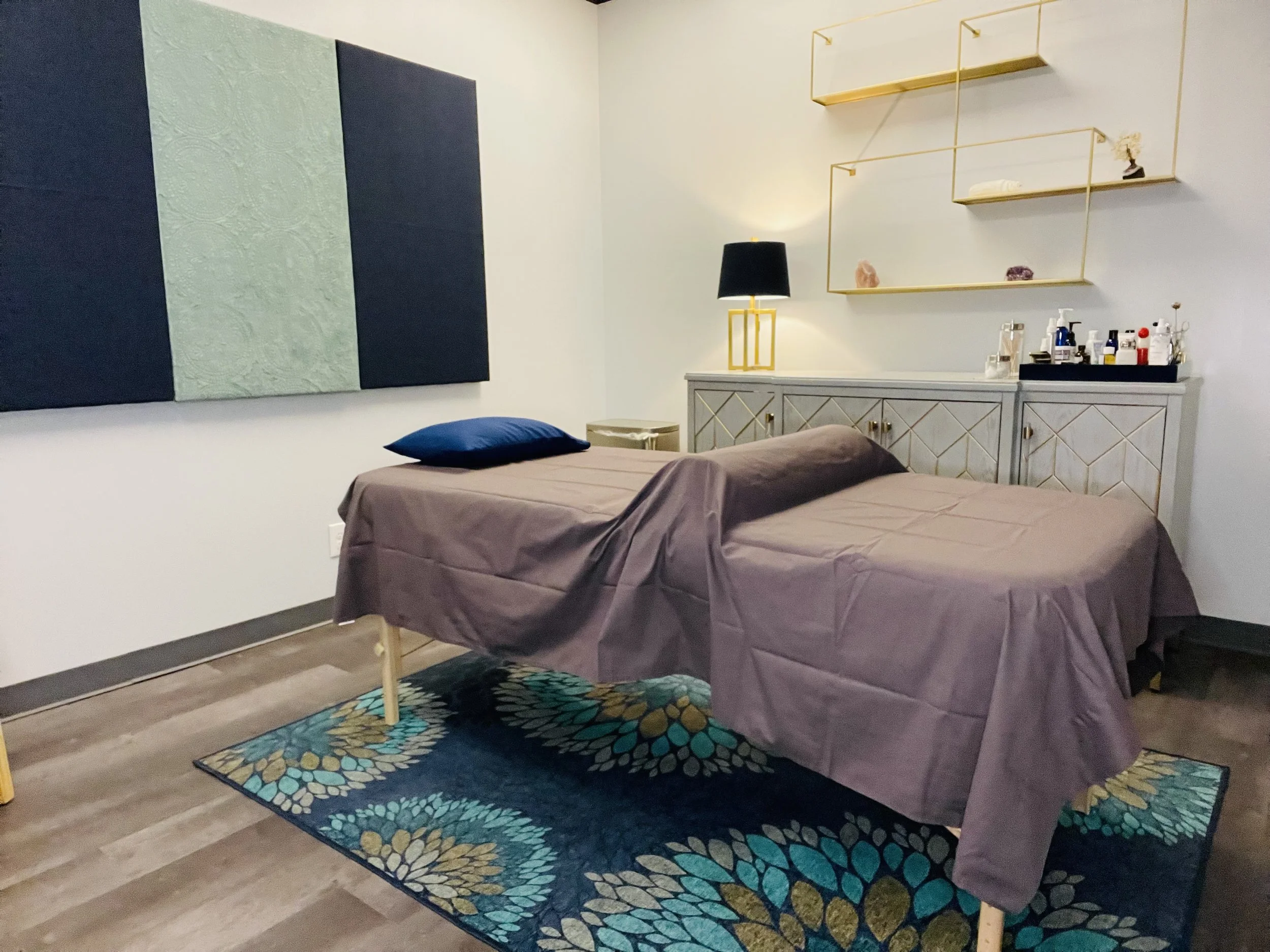 A bedroom with two beds covered in brown sheets and blue pillows, a colorful floral rug, a light gray dresser with gold handles, decorative shelves with rocks and a small statue, a black-lampshade table lamp, and a dark blue and green textured wall panel.