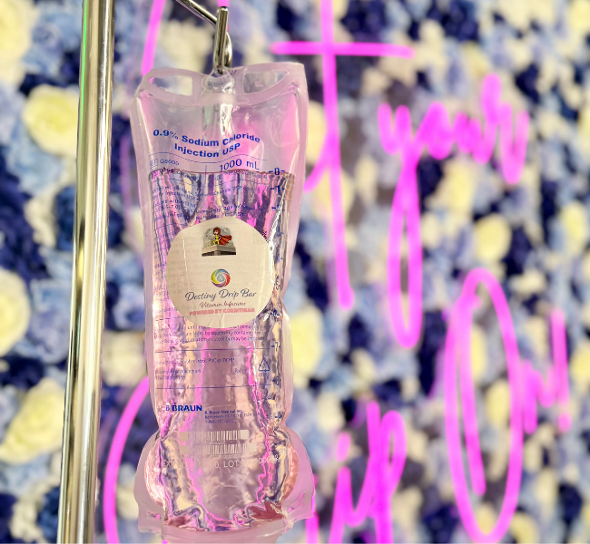 A clear intravenous drip bag filled with pink liquid hanging from a metal pole against a background of white flowers and a pink neon sign with handwritten text.