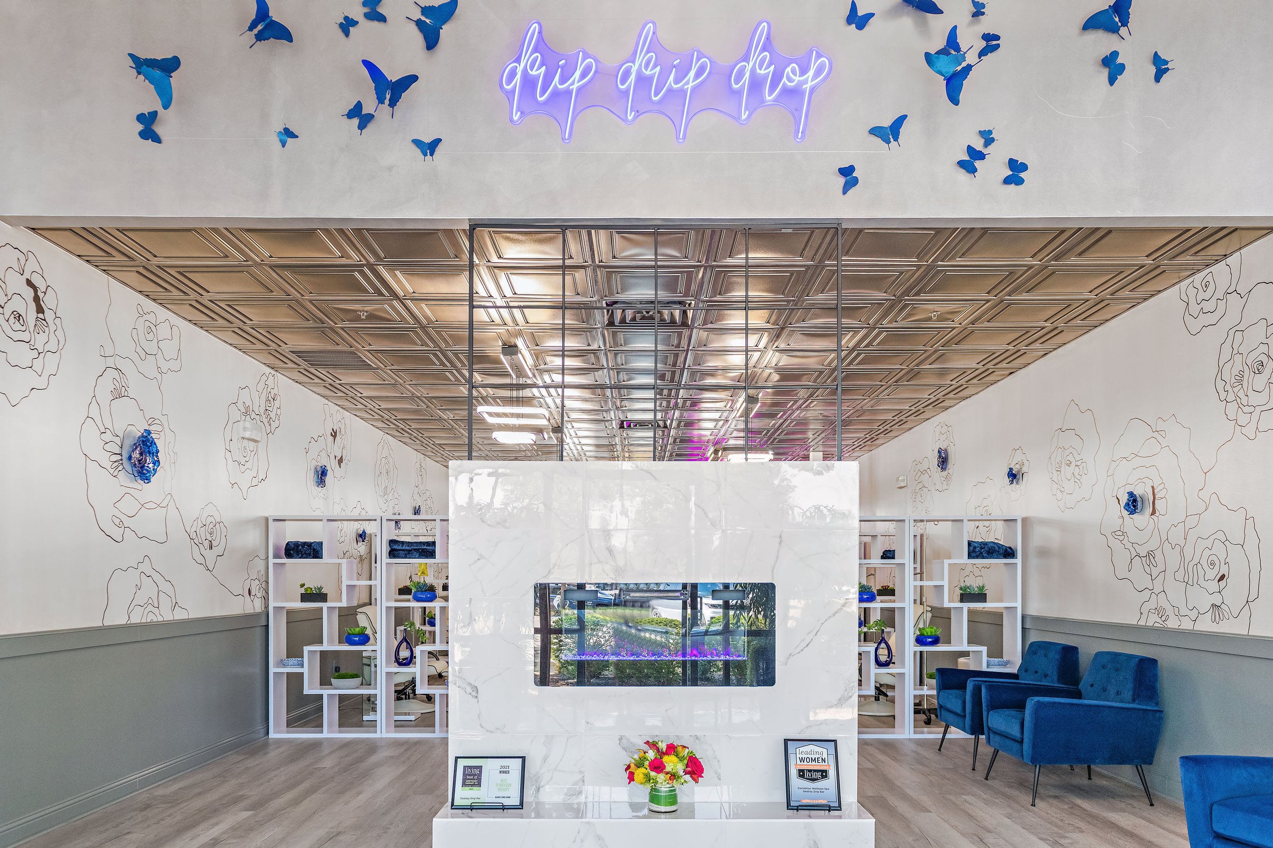Interior of a modern office or waiting area with white walls decorated with floral line art and blue flowers, a marble reception desk with framed certificates or awards, and blue seating on the right side. There are shelves with plants and decorative