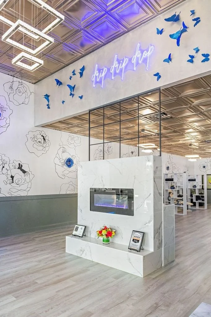 Modern interior space with a white marble fireplace, blue butterfly wall decorations, and a neon sign reading 'day drip drop.' There are magazines and a vase of flowers on the mantle, and geometric ceiling light fixtures.