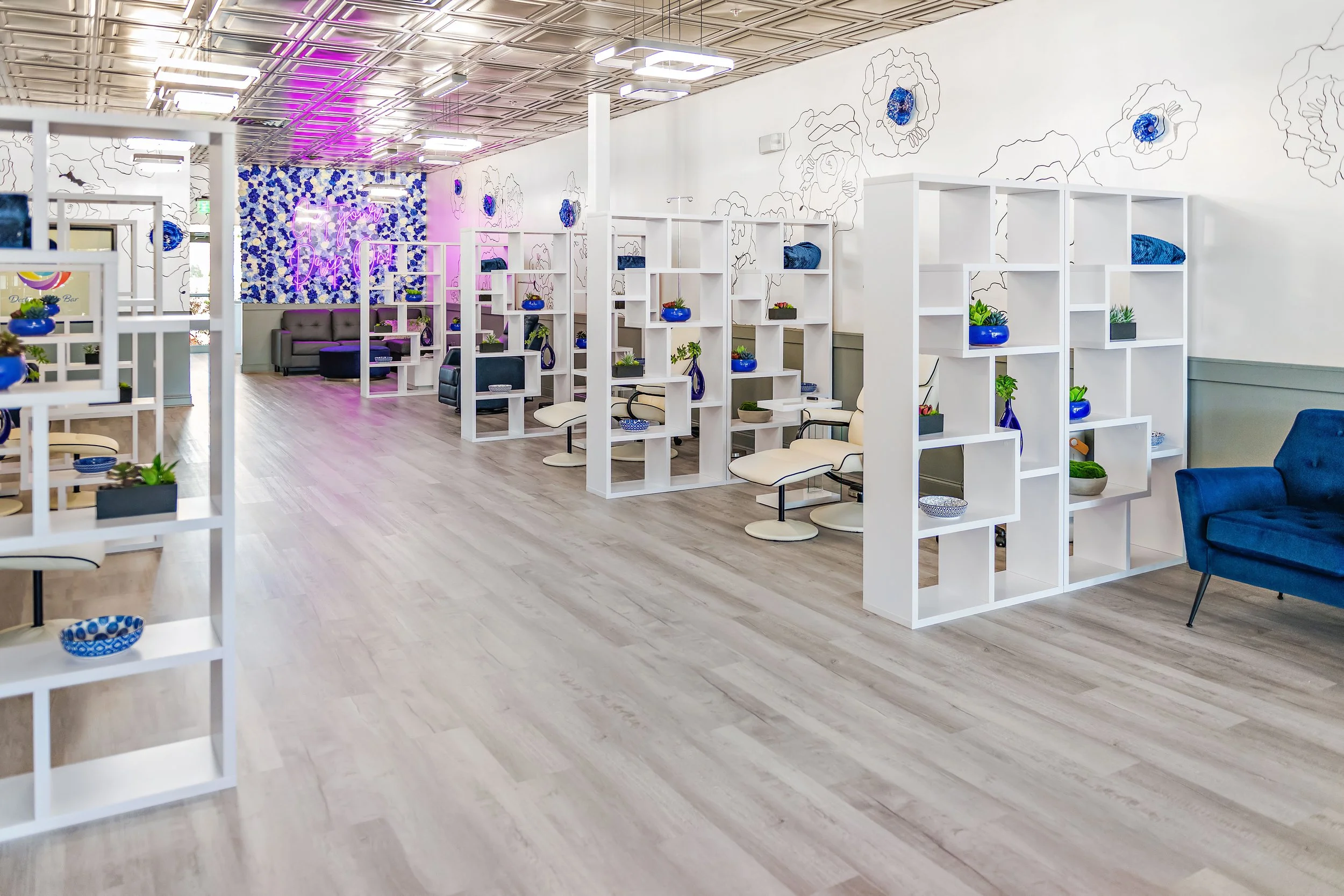 Modern, brightly lit interior space with white furniture and decorative shelving filled with blue vases and plants. The space features floral wall art, a neon sign, and a ceiling with metallic panels and geometric lighting fixtures.