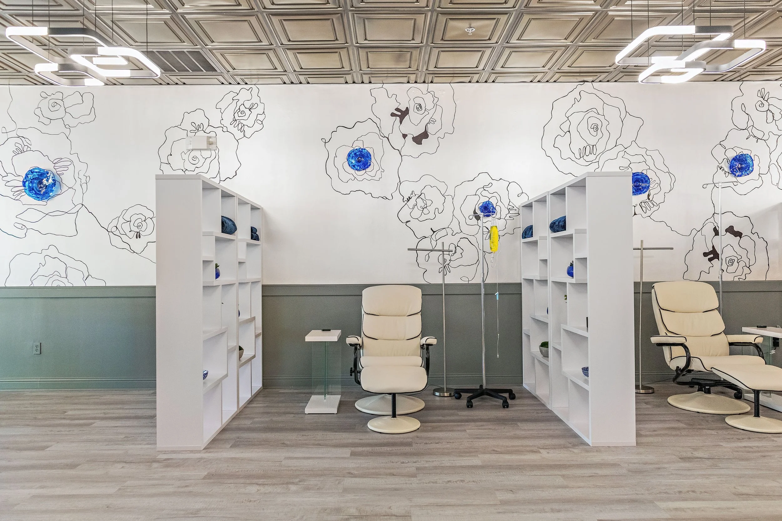A medical or office waiting room with beige chairs and white shelving units. A wall with floral line art and blue art pieces, ceiling with geometric light fixtures, and a wood floor.