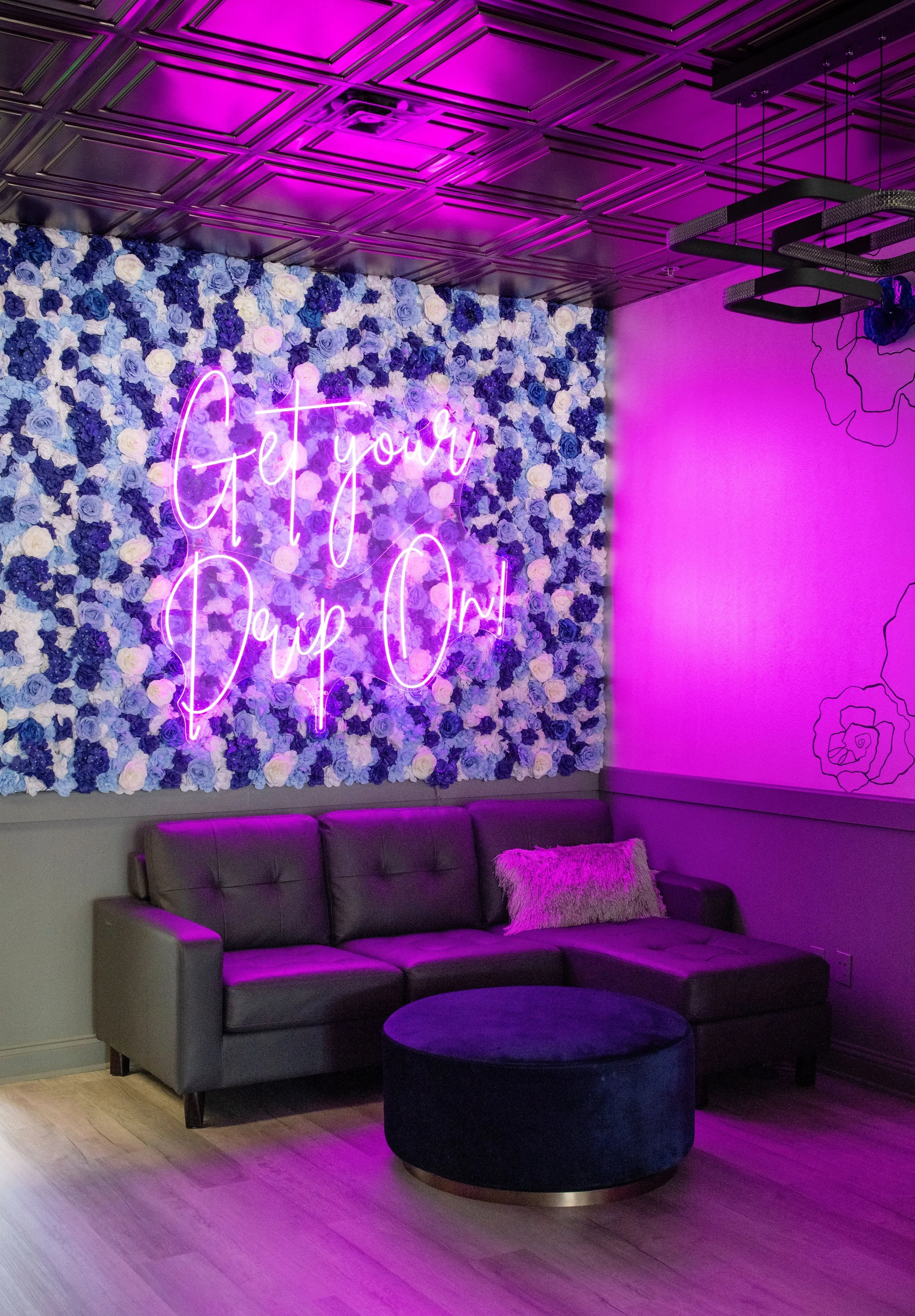 A cozy living room with a gray couch and a pink fluffy pillow. A round black ottoman is in front of the couch. The wall behind the couch is decorated with a wall of blue and white roses. A neon sign reading 'Get your Sip On!' glows in pink, mounted o