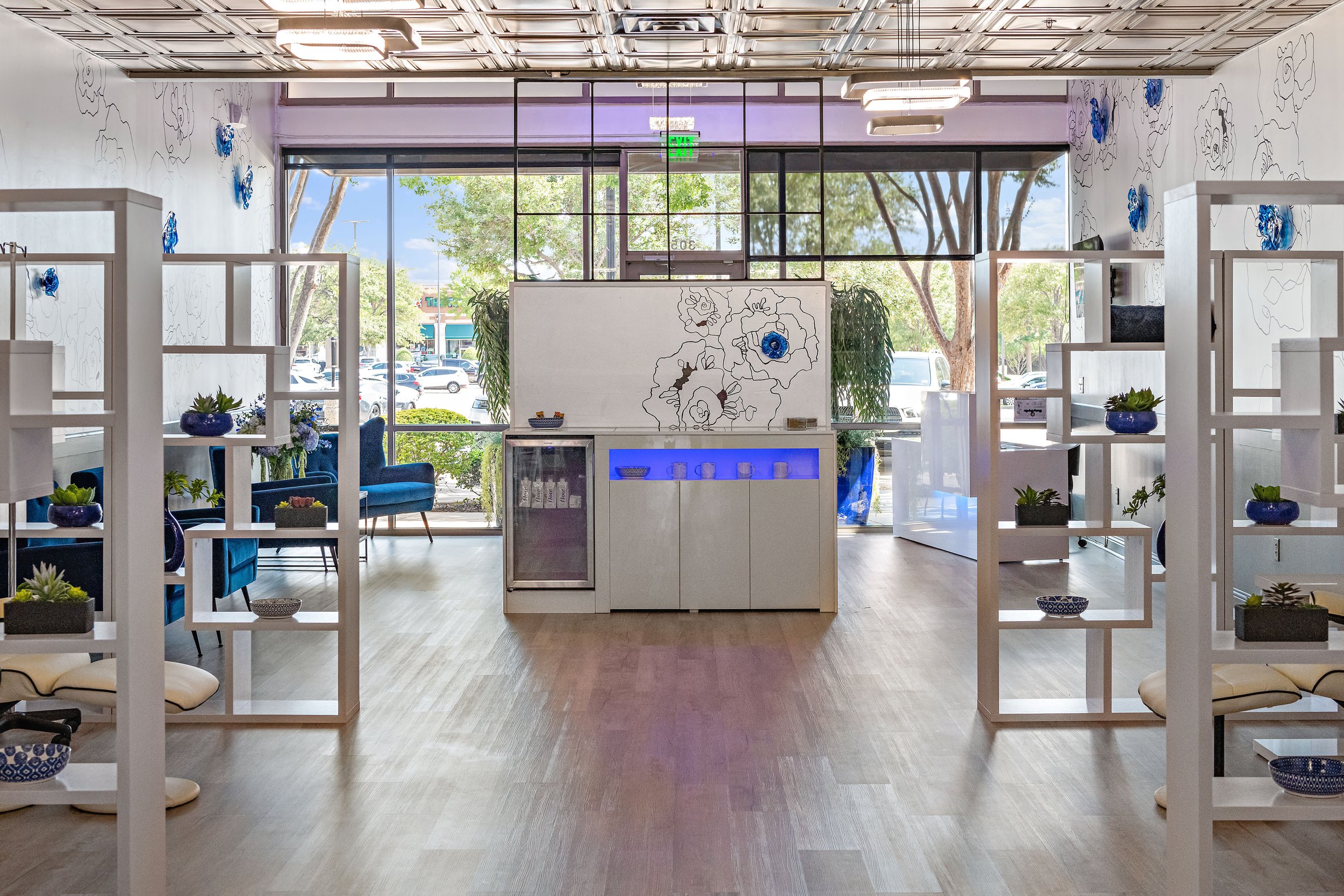 Interior of a modern, bright space with large windows showing trees and parking lot outside, featuring white shelving units with blue vases and plants, a white counter with a floral line drawing, and seating with blue chairs.