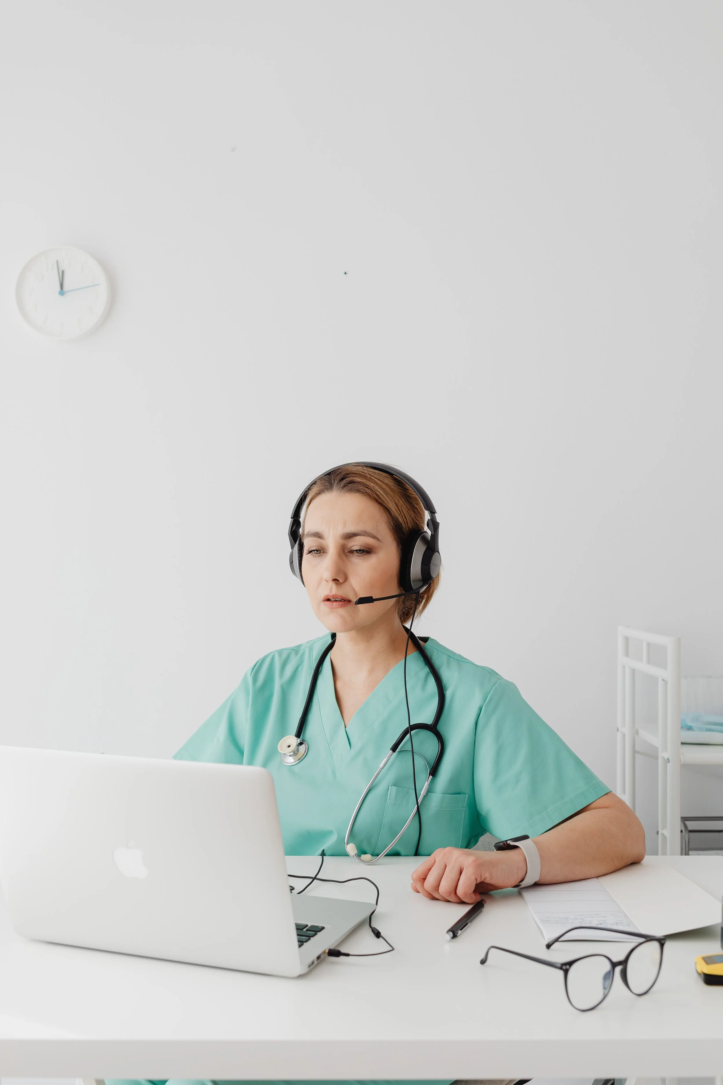 A female healthcare professional wearing green scrubs, using a laptop, with a stethoscope around her neck, and a smartwatch on her left wrist, seated at a white desk in a medical office.
