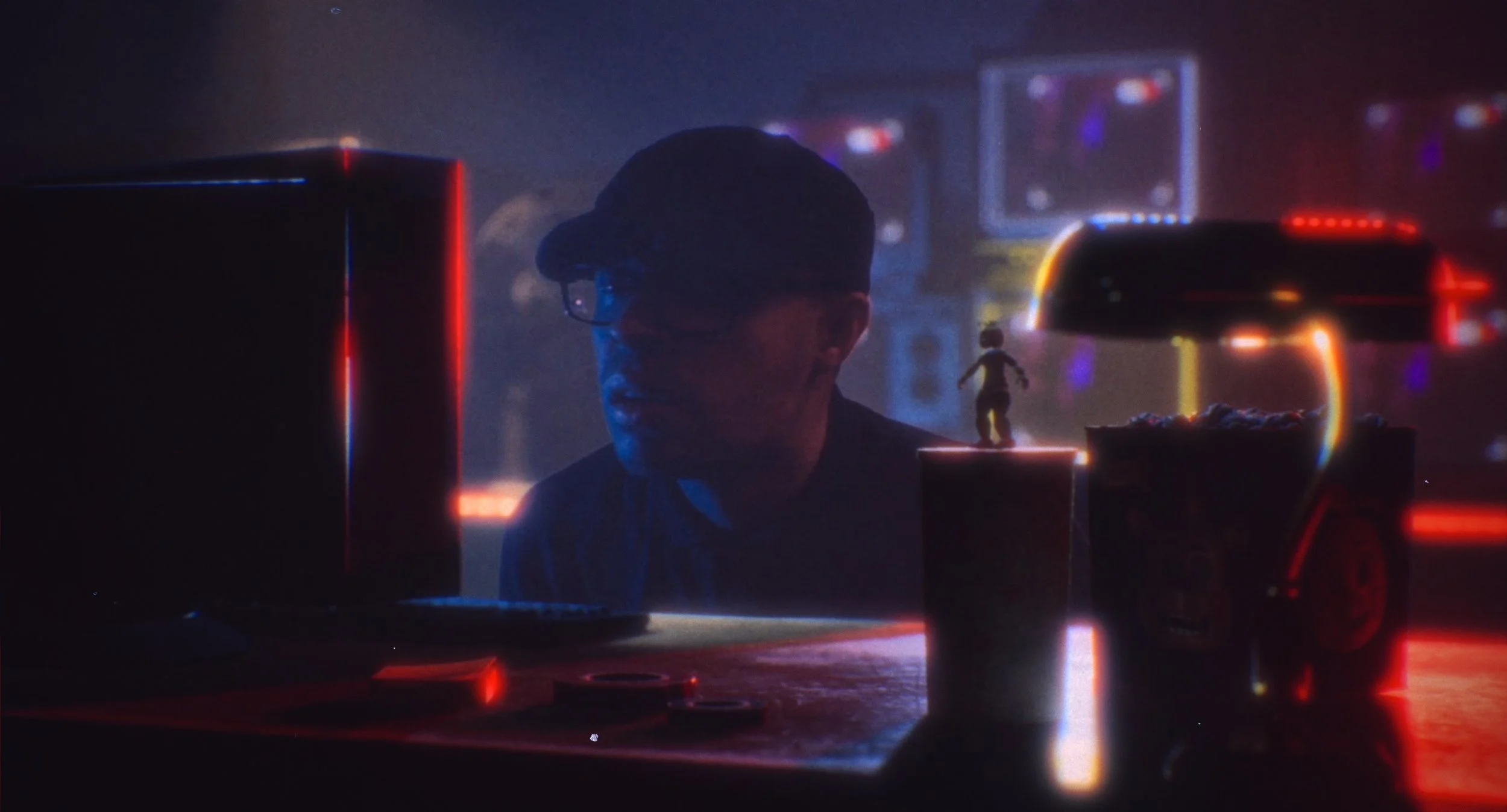 A person wearing glasses and a cap working on a computer with a small figurine on a shelf nearby, illuminated by neon red and blue lights in a dark room.