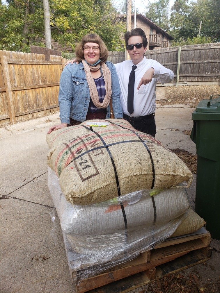  Rebecca and Jim smile and stand next to a shipment of cacao on a pallet behind Cultiva Labs. Jim points at the cacao. 