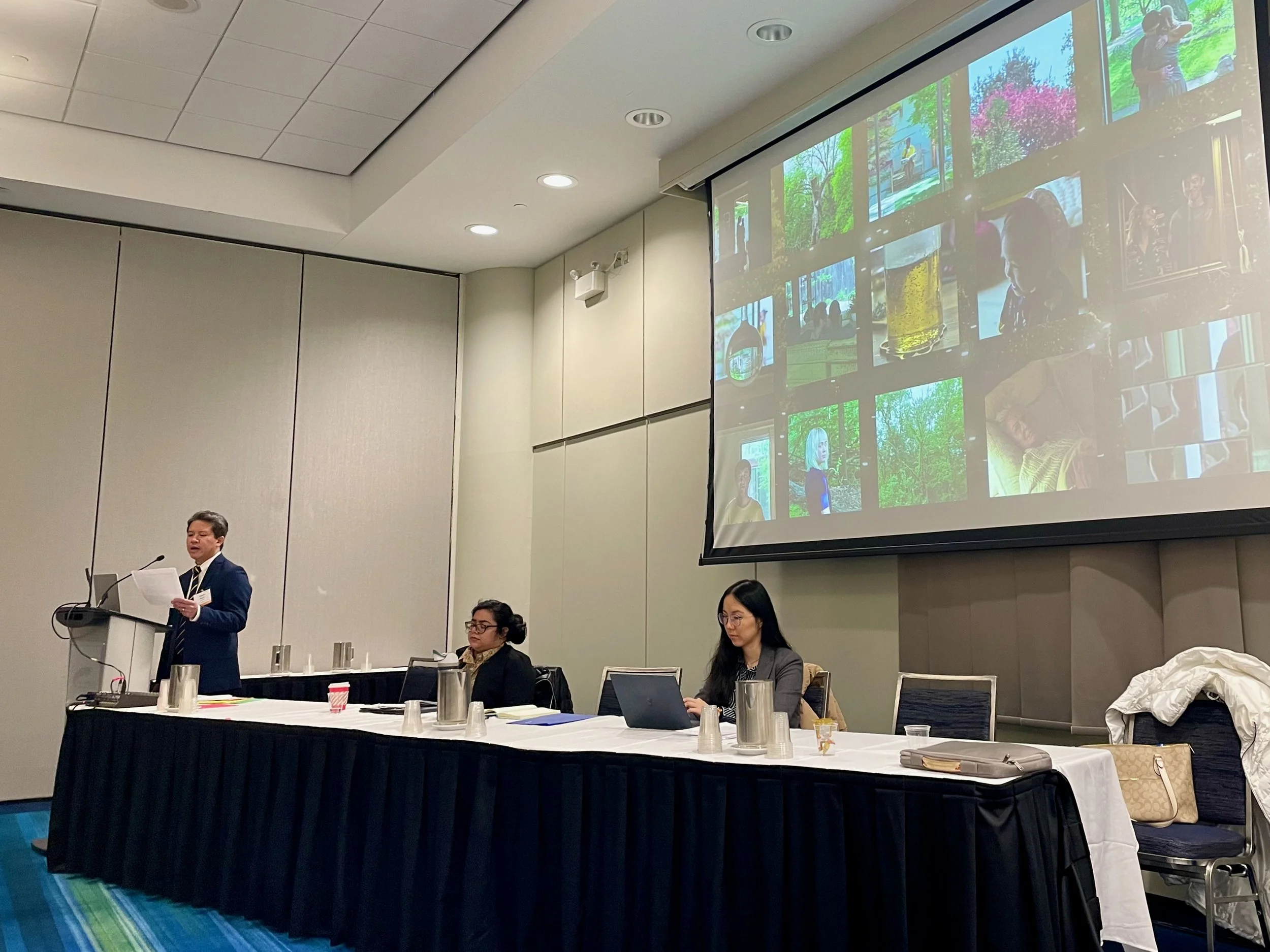 Academic conference panel. Presenter reading at podium. Two persons behind the front table. Screen behind w/ collage of still images from film