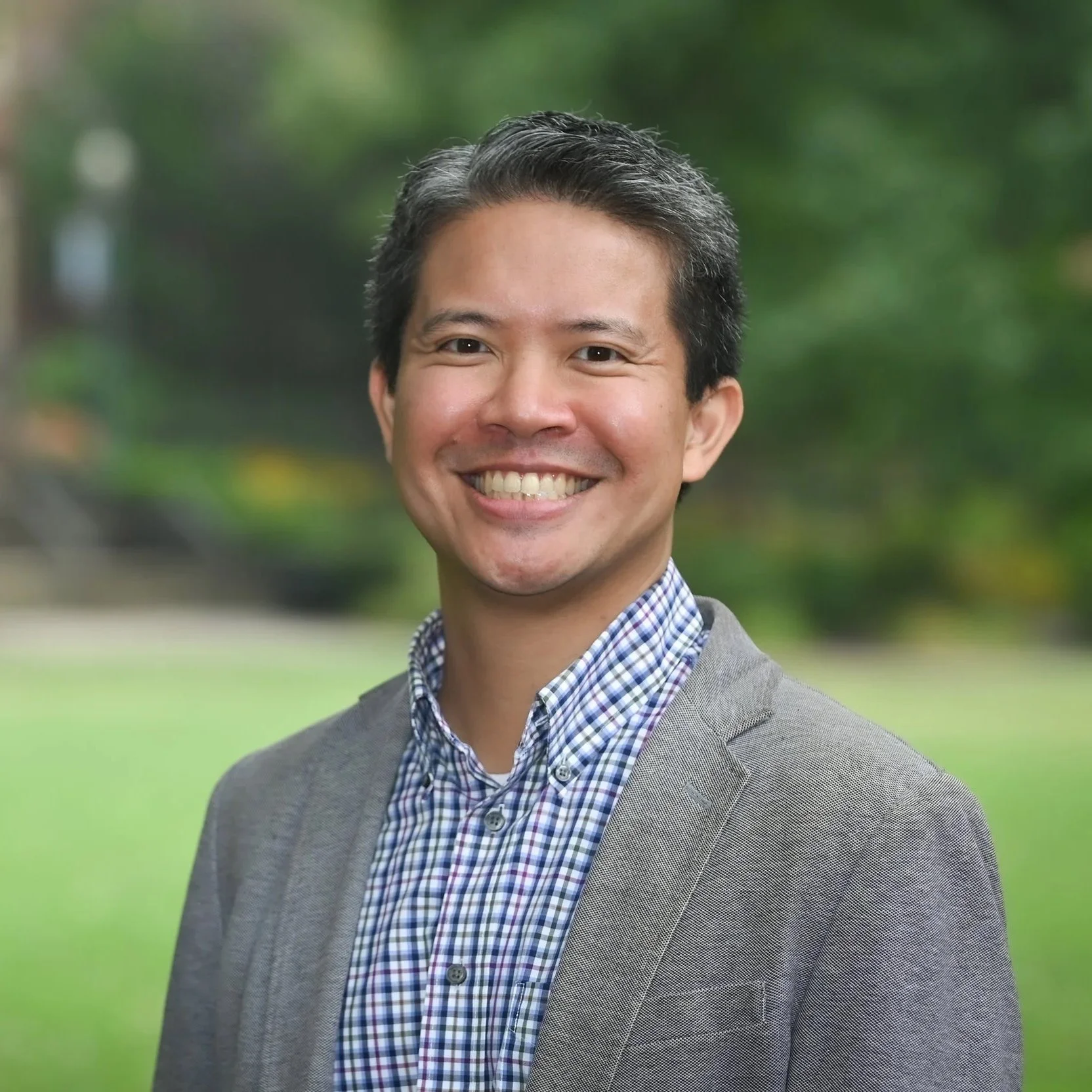 Middle-aged Asian male wearing gingham shirt and blazer. Blurred campus background.