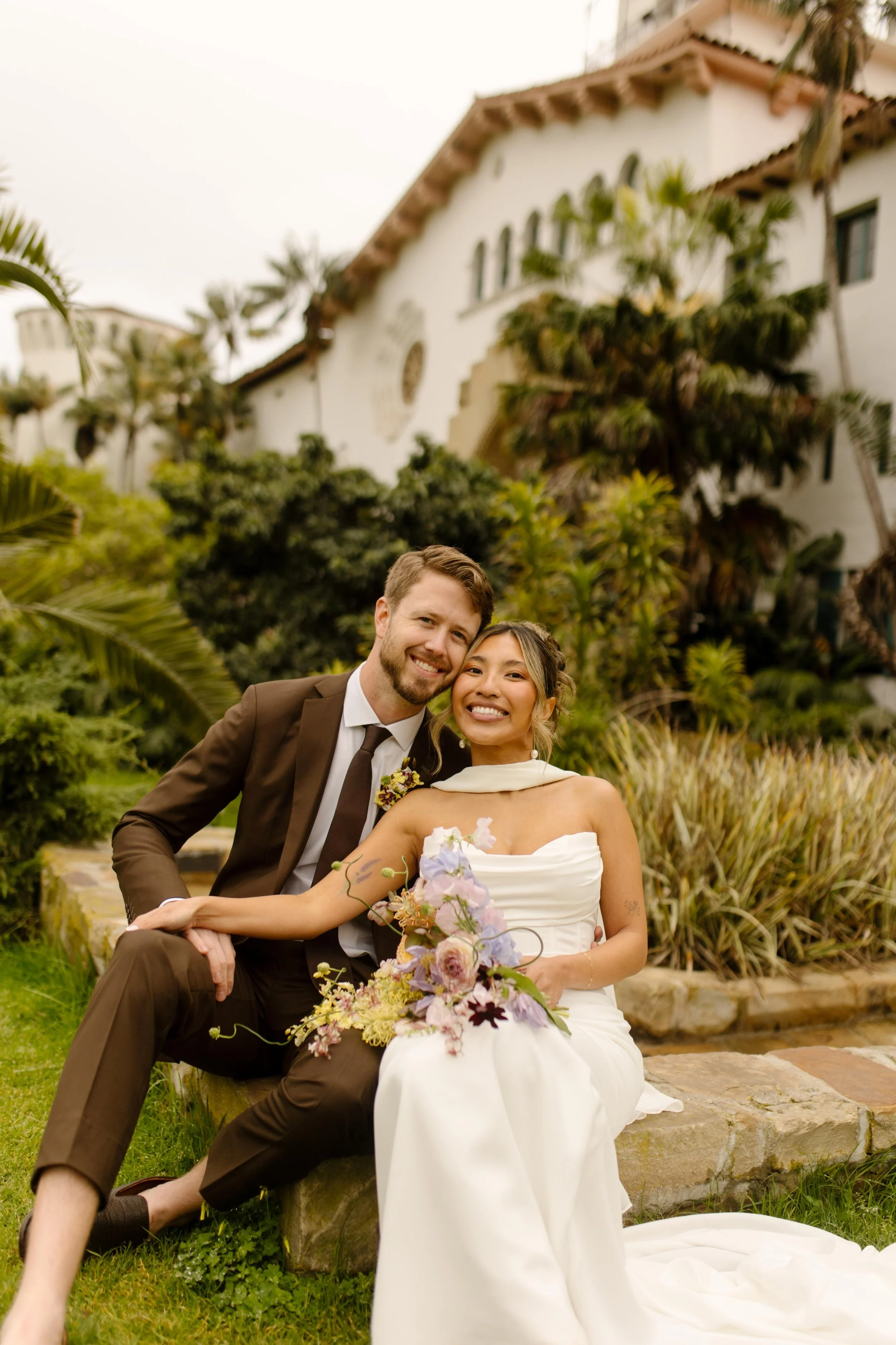 A joyful couple sits on a stone bench surrounded by lush greenery, smiling wide in front of the historic architecture of their wedding Santa Barbara Courthouse.