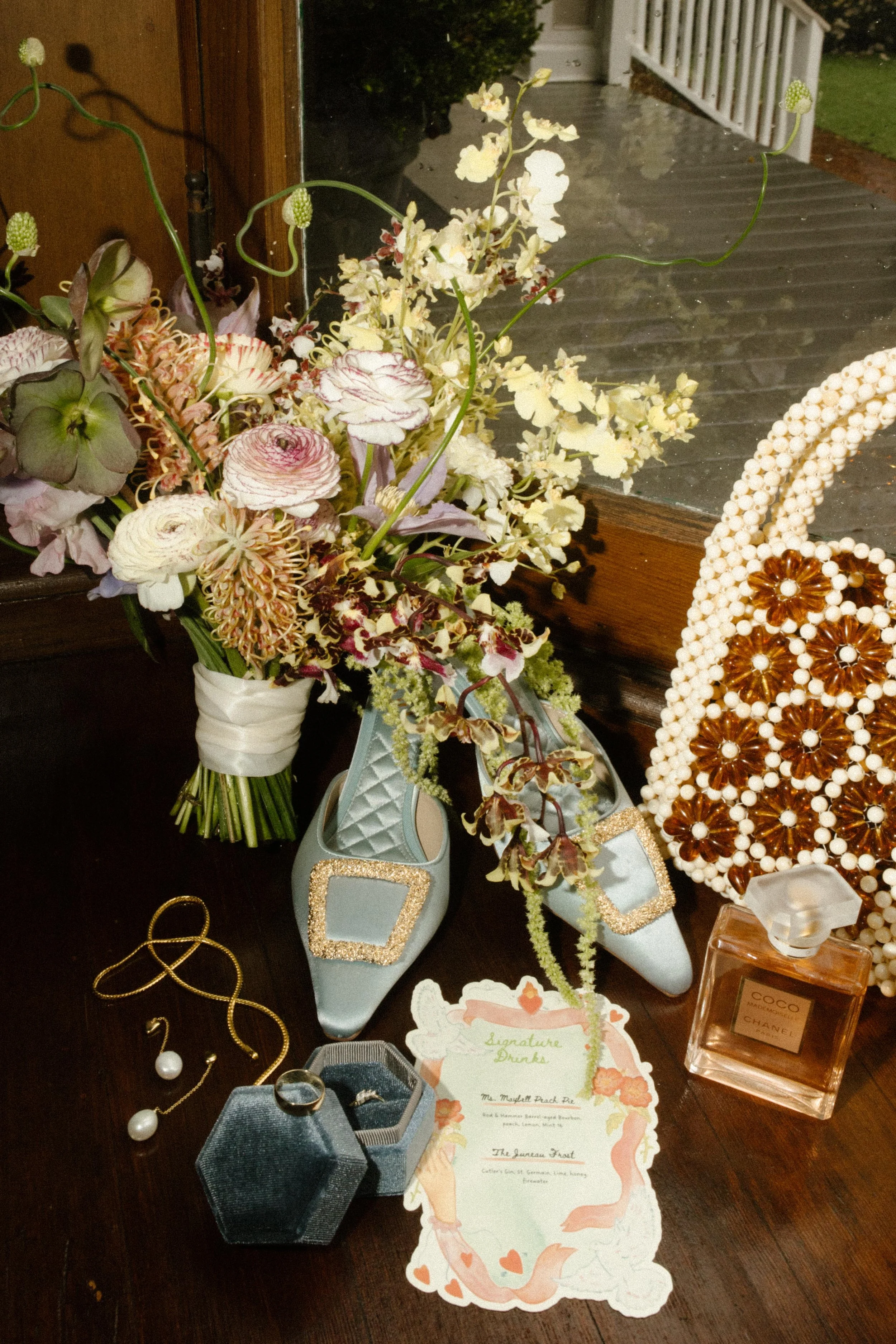 Flat lay of the bride’s details: soft blue shoes, a floral bouquet, vintage-inspired jewelry, and a wedding invitation styled on a wooden windowsill.
