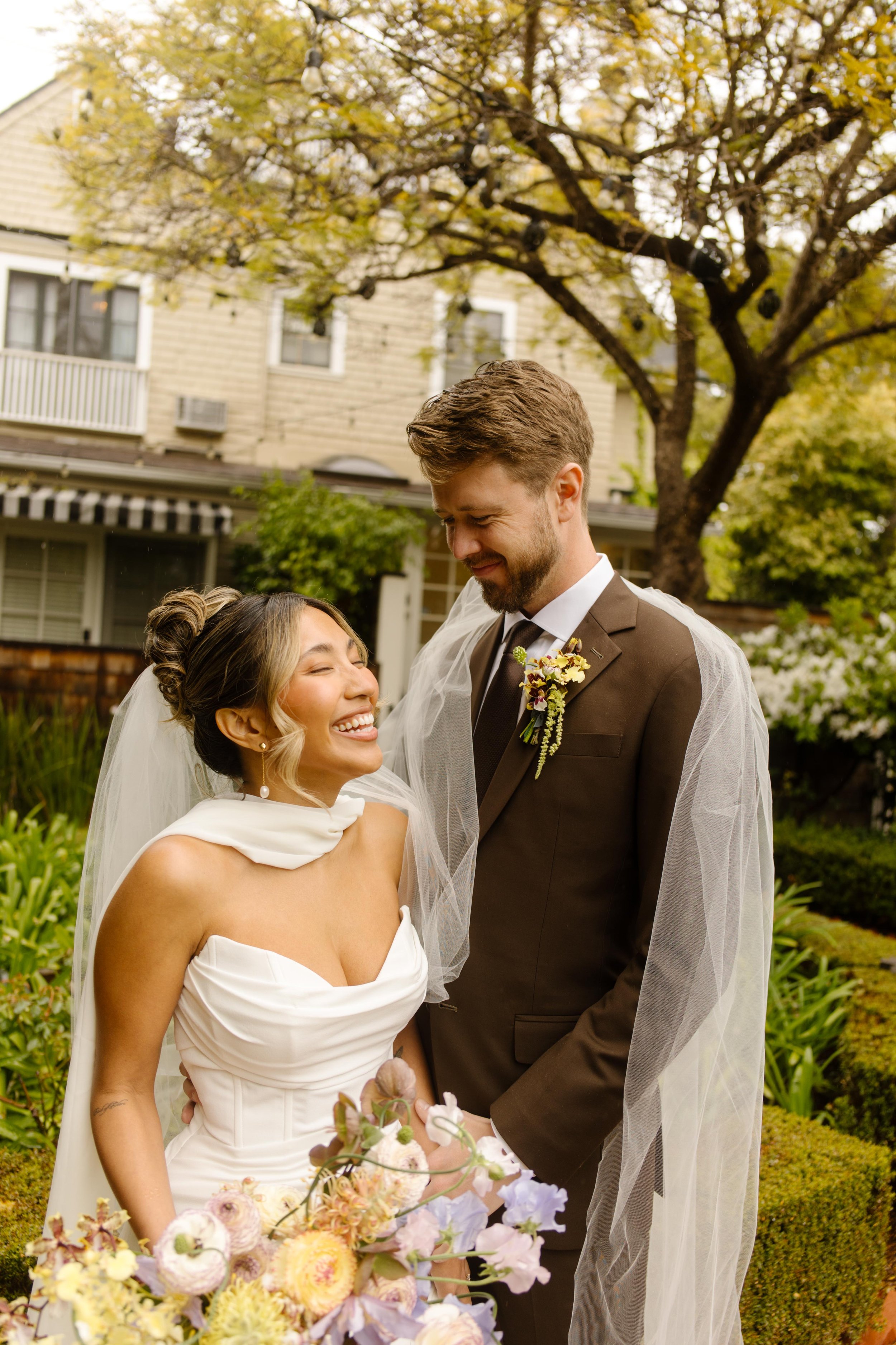 The bride laughs with her groom in a garden courtyard, holding a bouquet of pastel blooms as they share a private moment.
