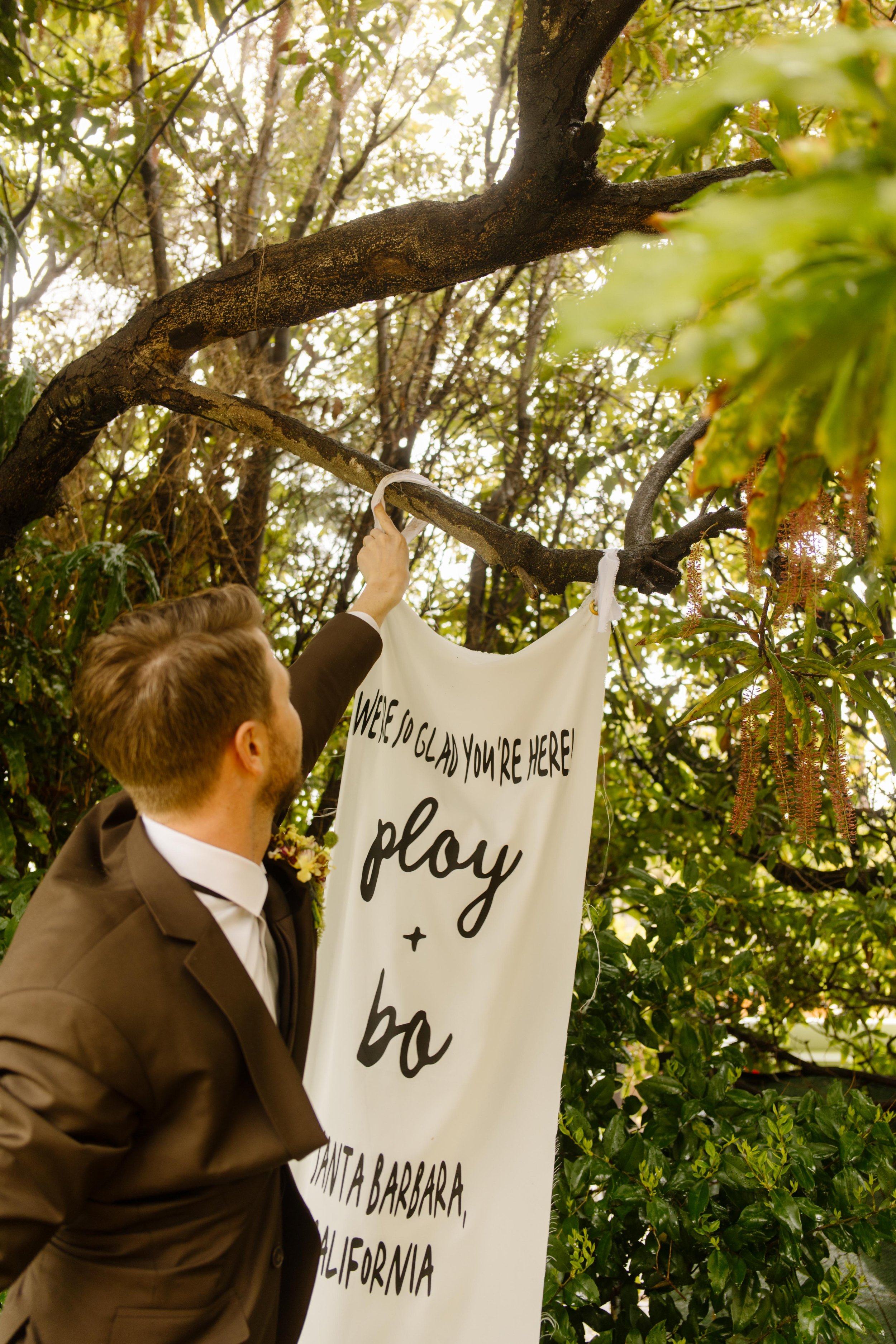 The groom hangs a welcome banner that reads “Ploy + Bo, Santa Barbara, California,” beneath a shady tree.