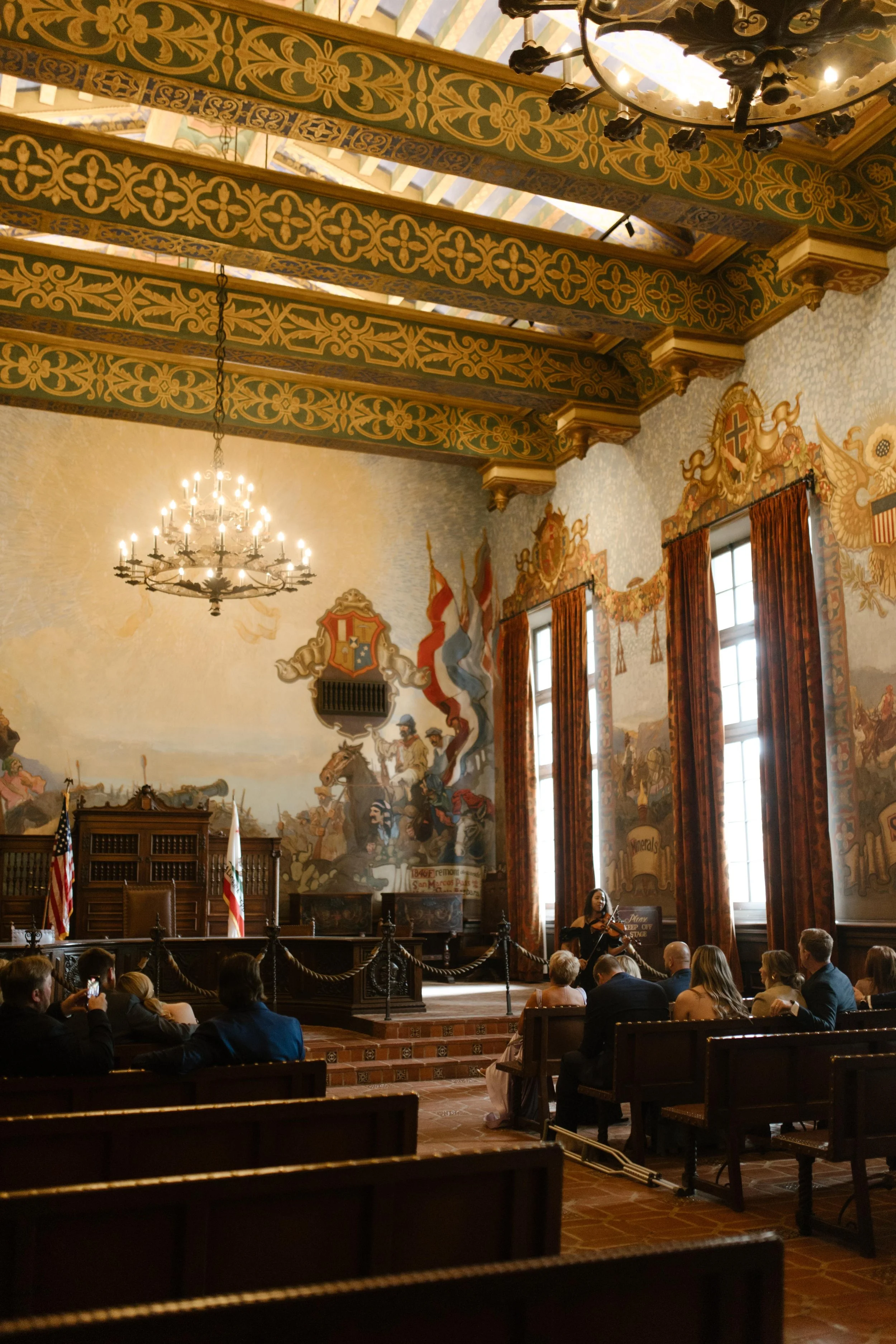 Guests sit beneath the grand painted ceiling of the mural room during a wedding Santa Barbara Courthouse ceremony, surrounded by golden light and historic detail.